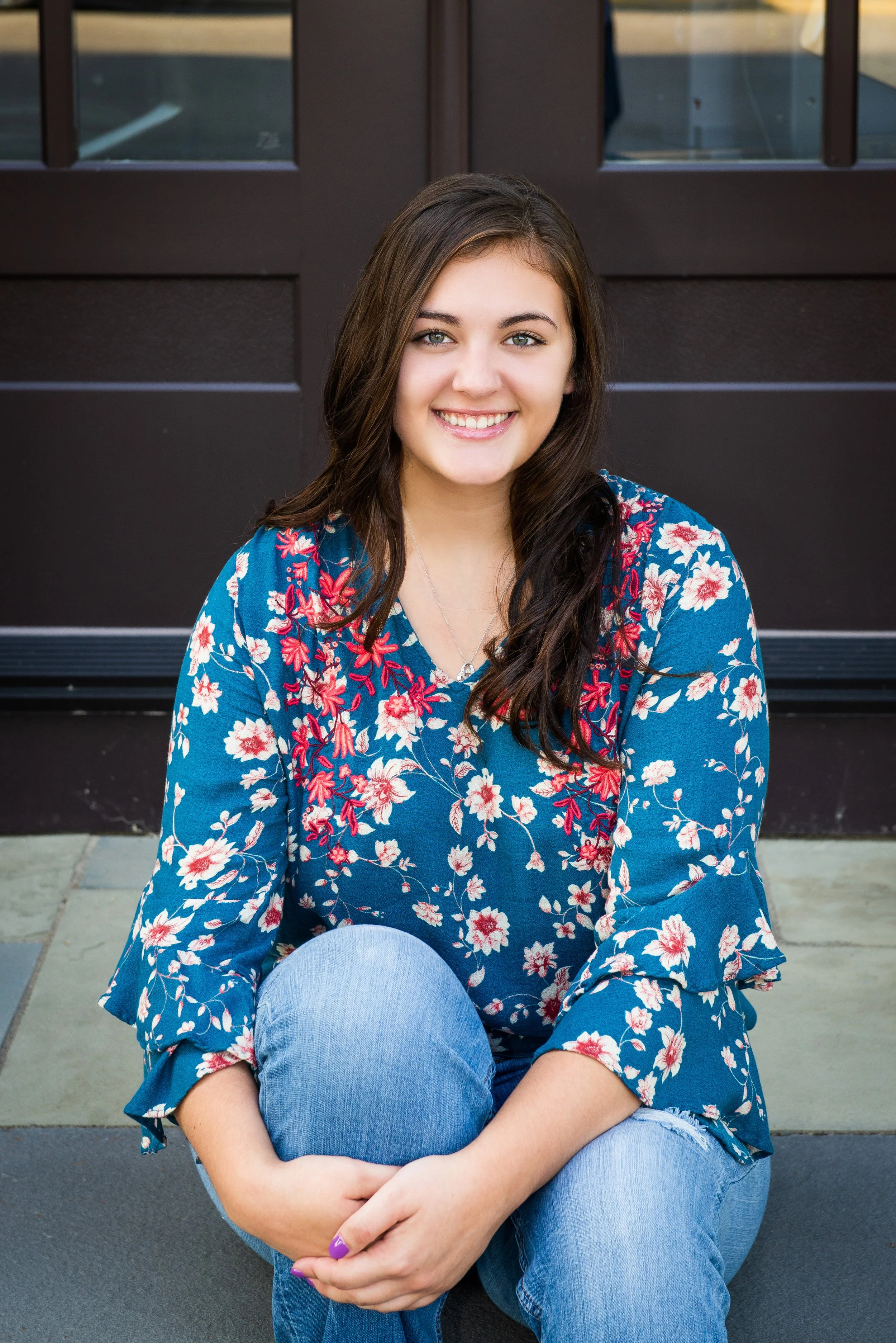 A young woman with long dark hair, blue eyes, and fair skin, wearing a blue floral top and blue jeans, sitting outdoors on paved ground in front of a dark brown door with glass panels.