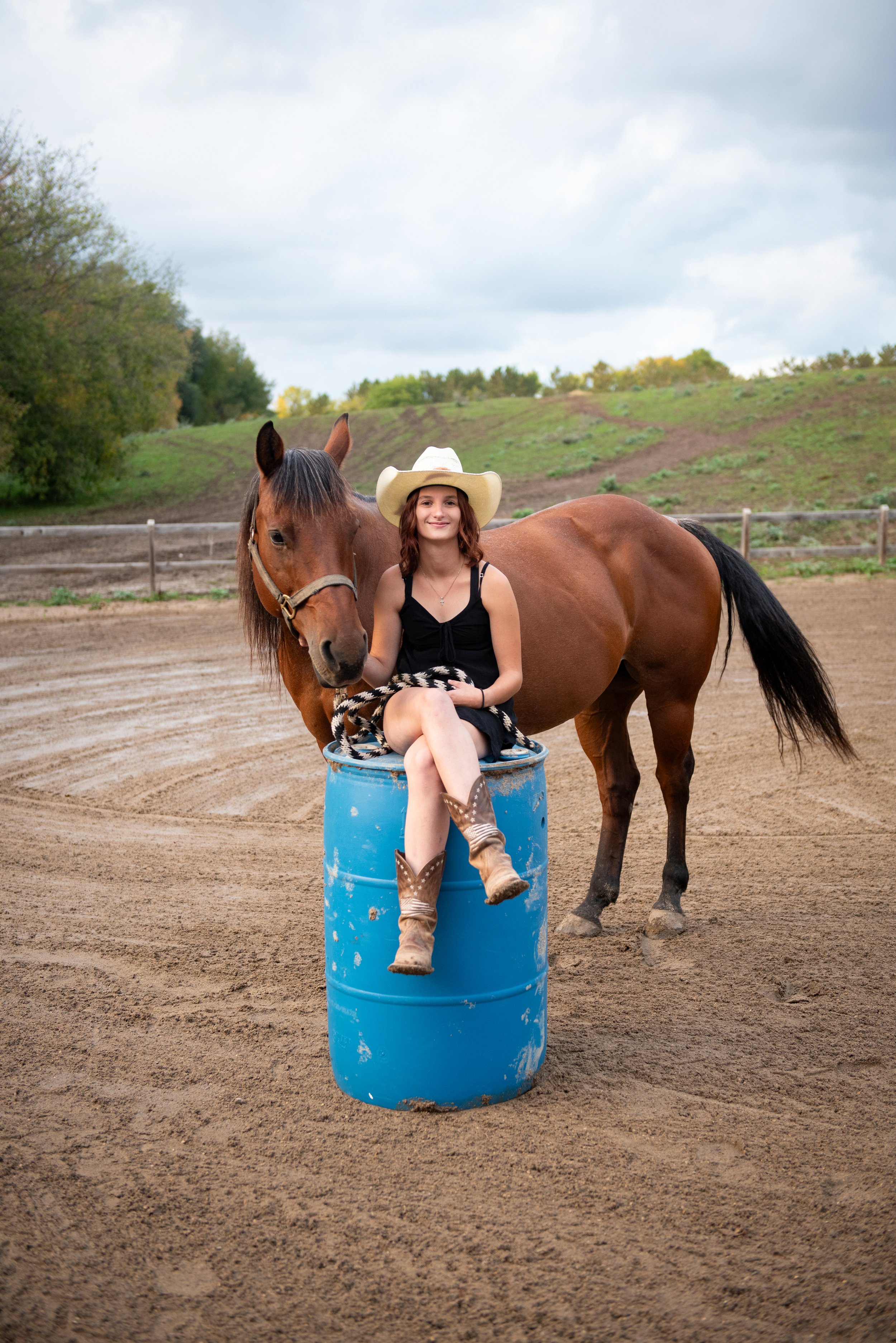 A young woman in cowboy boots and a black dress, wearing a cowboy hat, sitting on a blue barrel, smiling, with a brown horse standing beside her on a dirt rodeo or ranch arena.