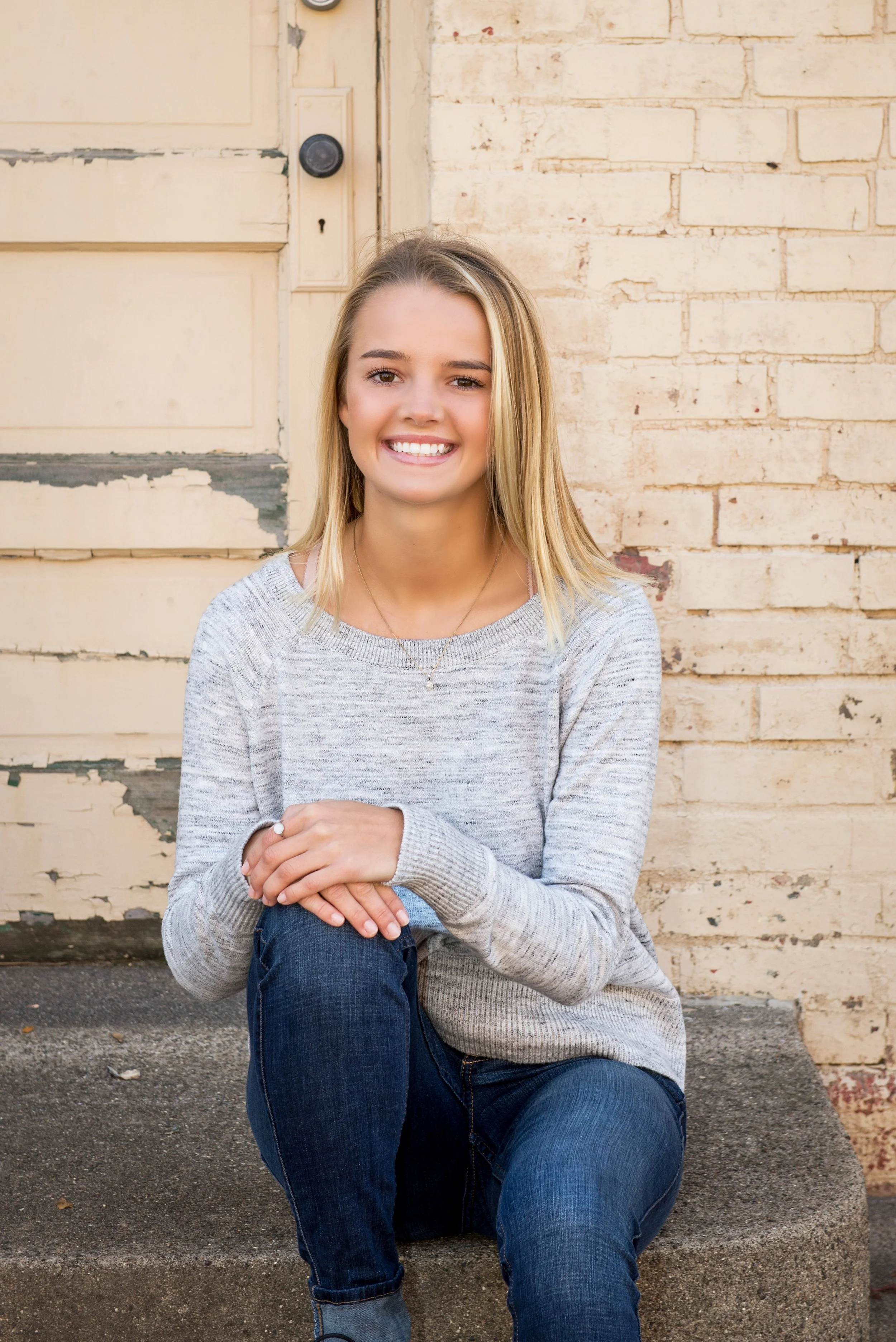 A young woman with blonde hair and a gray sweater smiling while sitting on a sidewalk in front of a brick wall and old door.