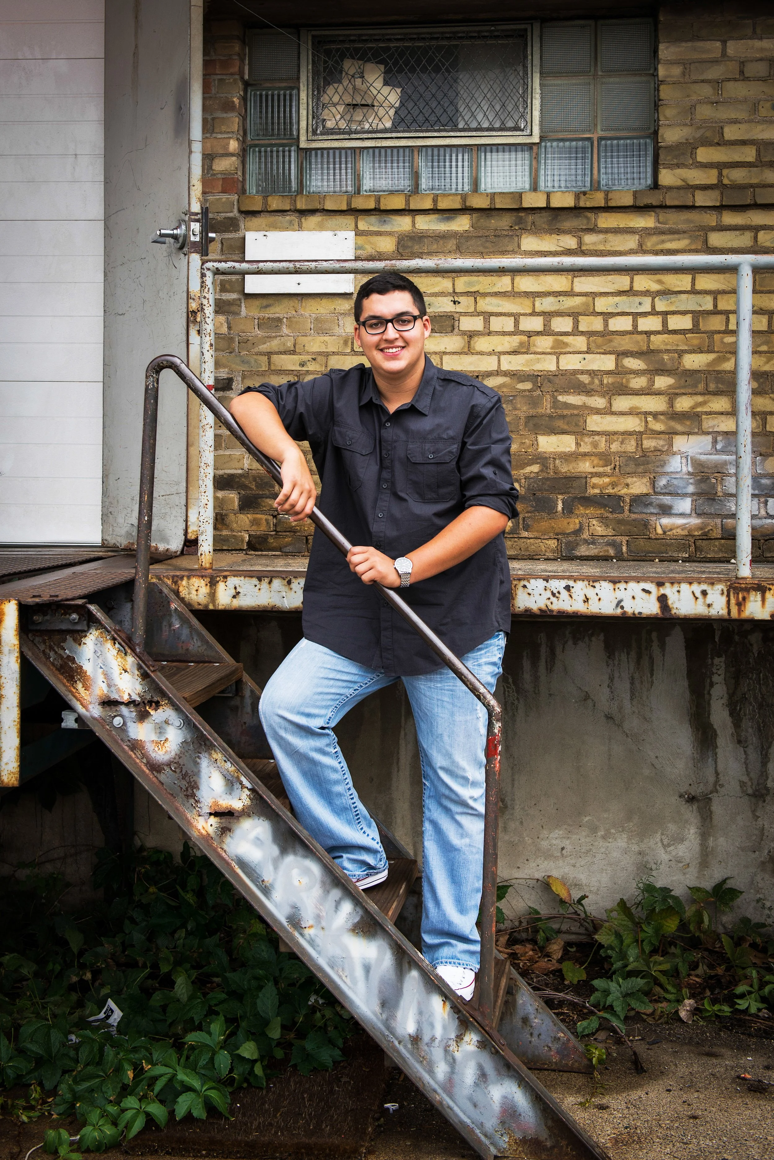 A young man with glasses, short dark hair, and a friendly smile, standing on a rusty metal staircase outside a brick building, wearing a black button-up shirt, light blue jeans, and white sneakers.
