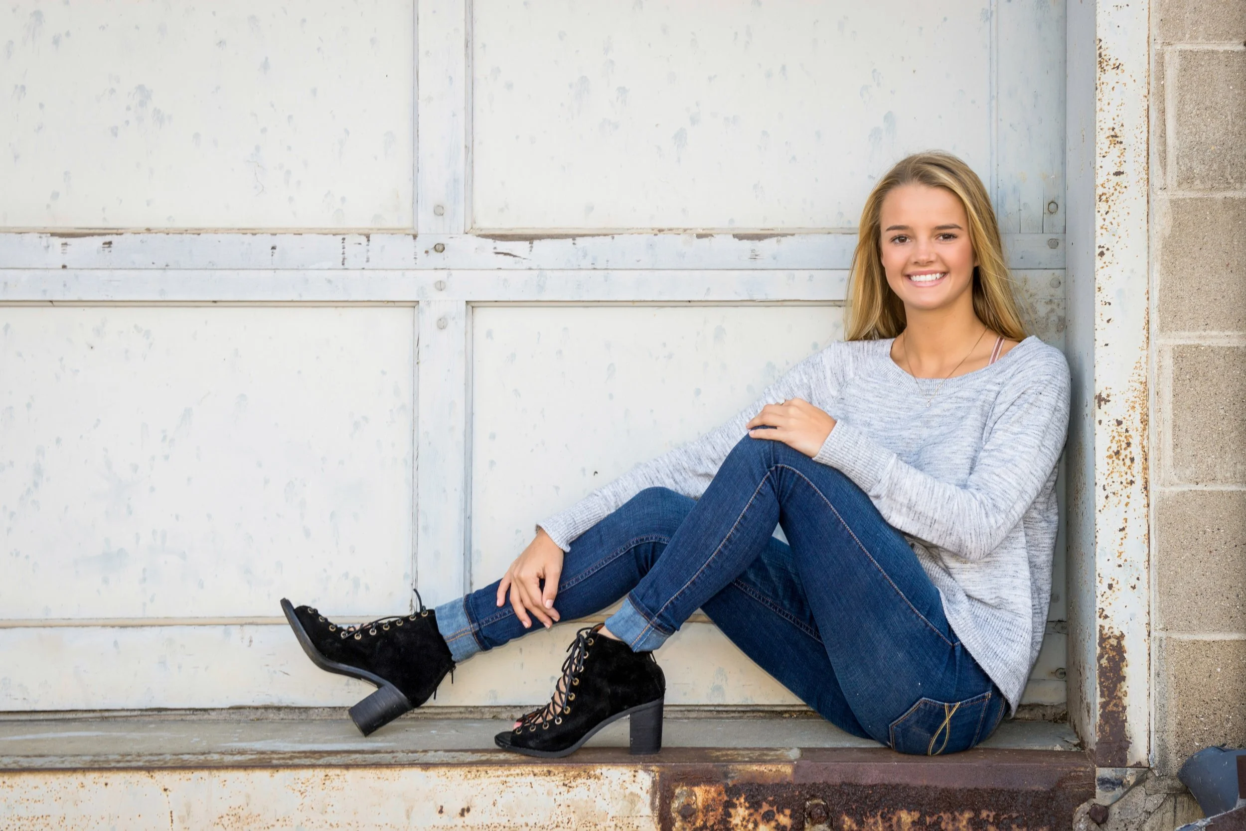 A young woman with long blonde hair sitting on the ground, smiling, against a white garage door with a concrete block wall on the right, wearing a gray sweater, blue jeans, and black heeled boots.