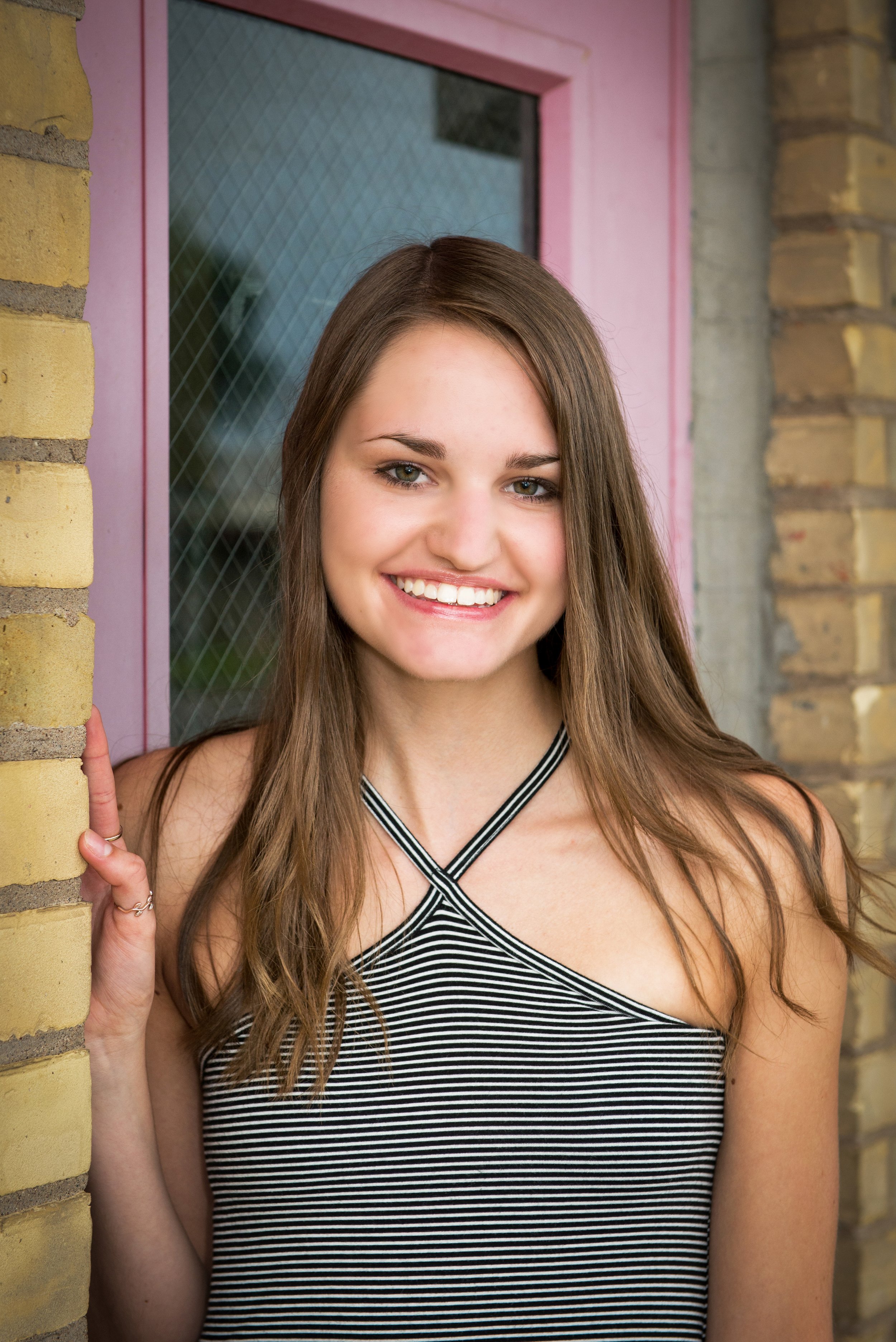 A young woman with long brown hair smiling, holding a brick wall, standing in front of a pink door with a glass window.