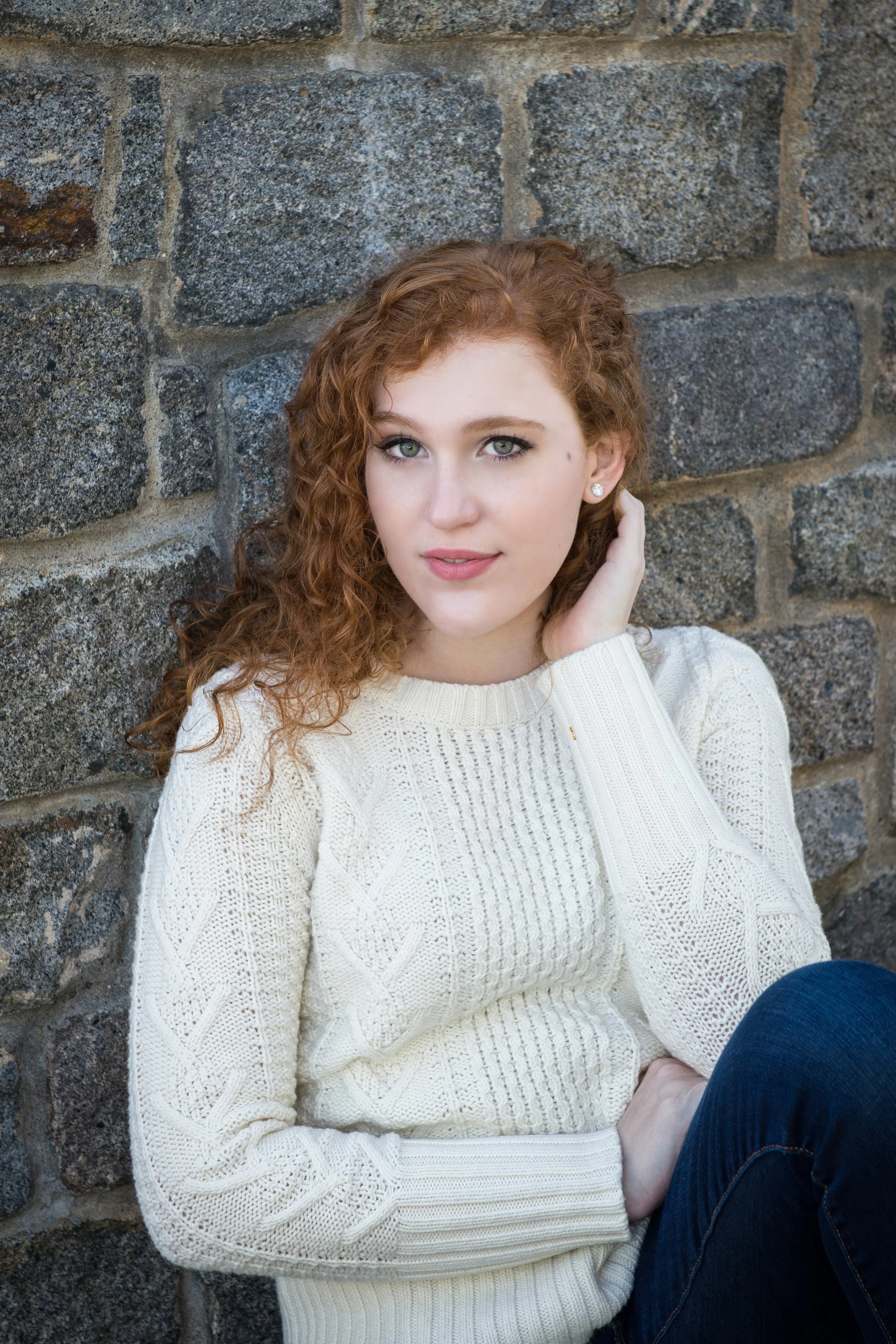A young woman with red curly hair and blue eyes, wearing a white knitted sweater and dark jeans, sitting against a stone wall.