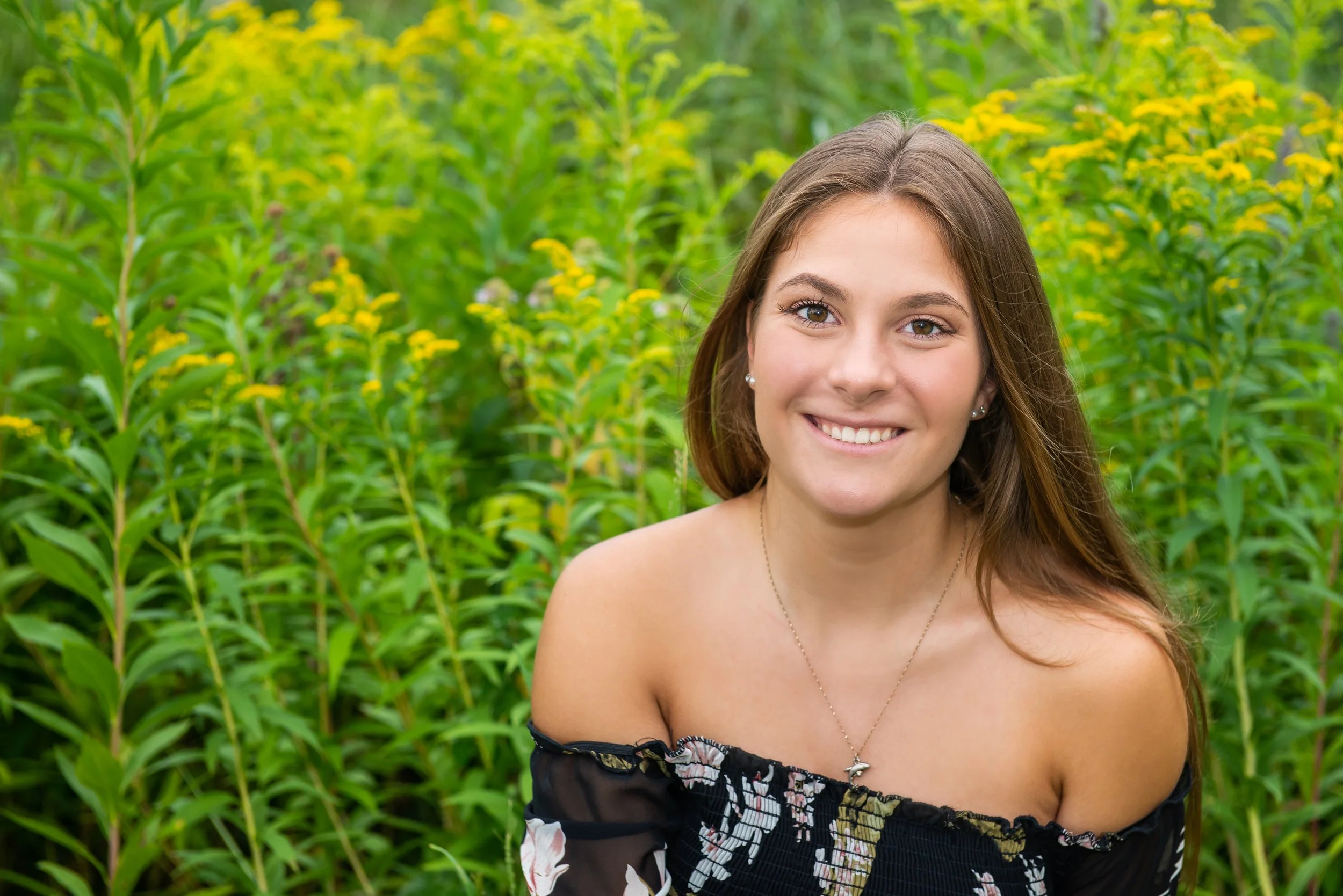 A young woman with long brown hair smiling outdoors in a green, yellow-flowered field, wearing an off-the-shoulder black floral top and a necklace.