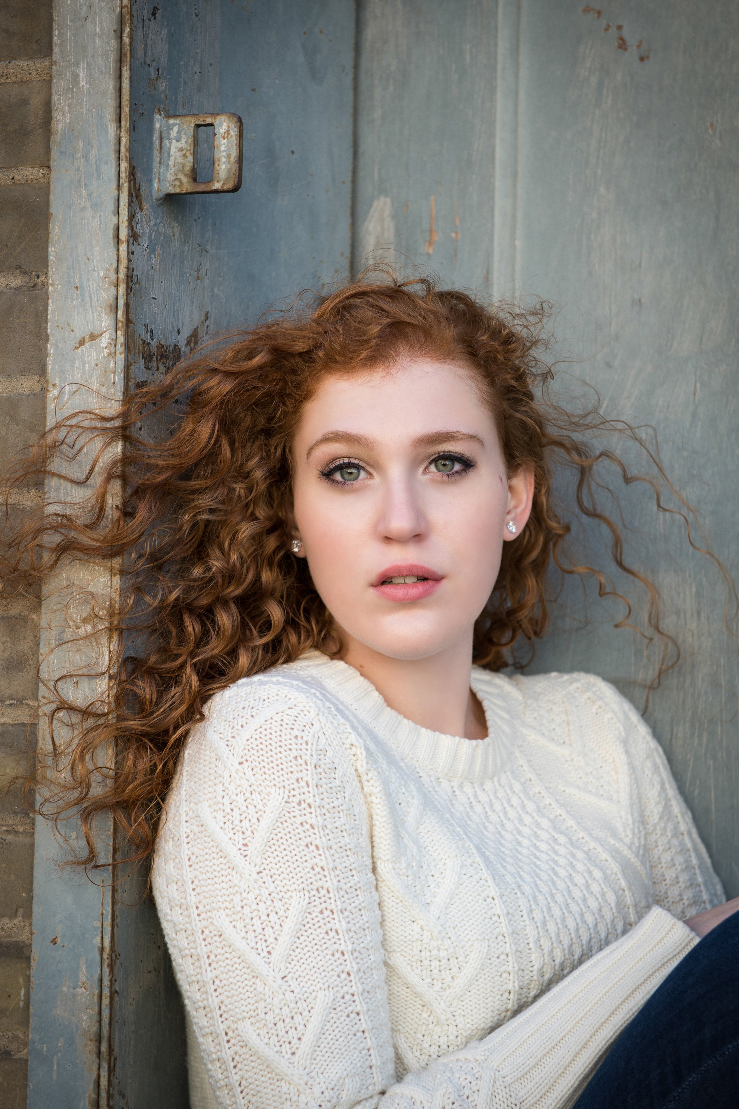 A young woman with curly red hair and green eyes, wearing a white knitted sweater, sitting against a weathered blue wall with a brick corner, looking directly at the camera.