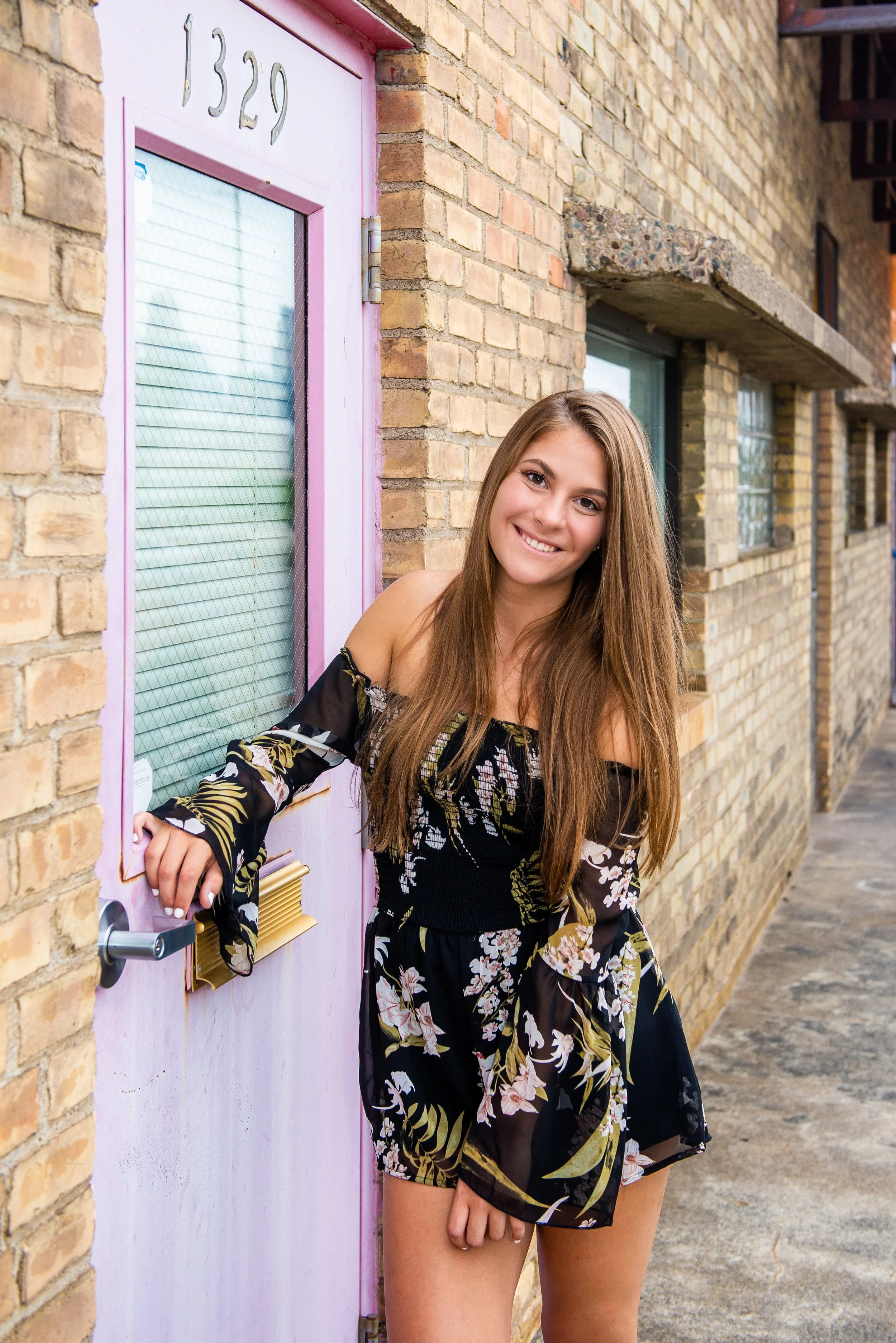 A young woman with long brown hair, wearing a black floral off-shoulder dress, smiling while leaning on a pink door with the number 1329, in front of a brick building.