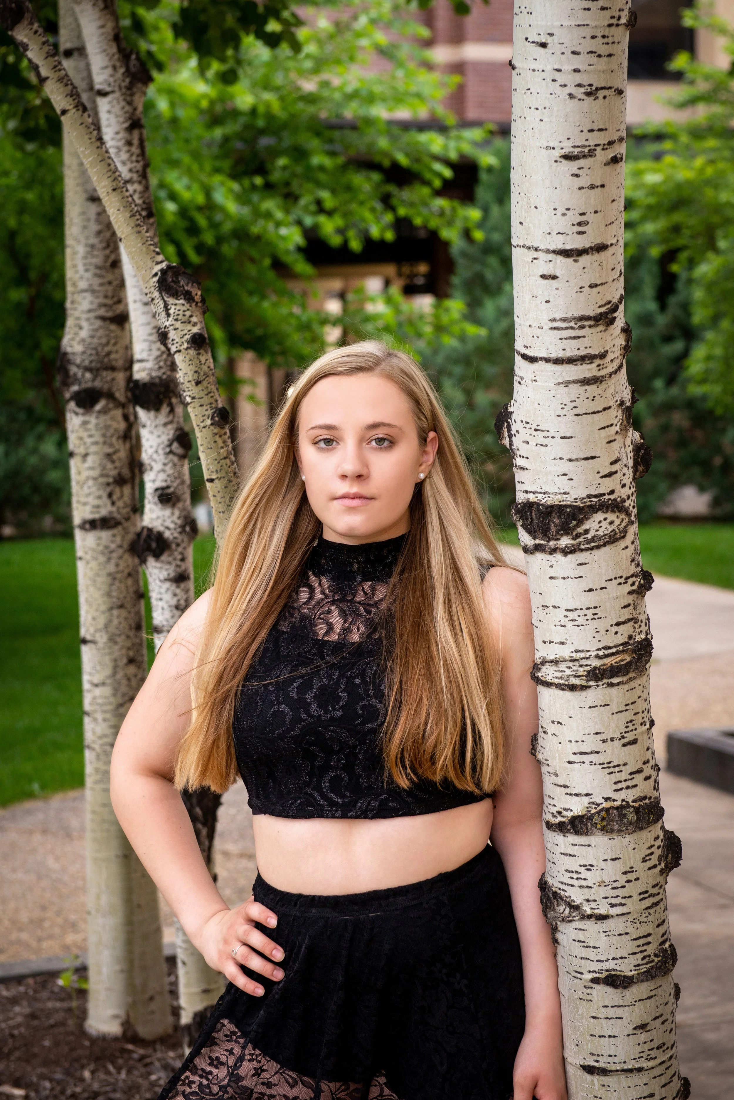 A young woman with long blonde hair wearing a black lace crop top and skirt, standing outdoors next to a birch tree with green foliage in the background.