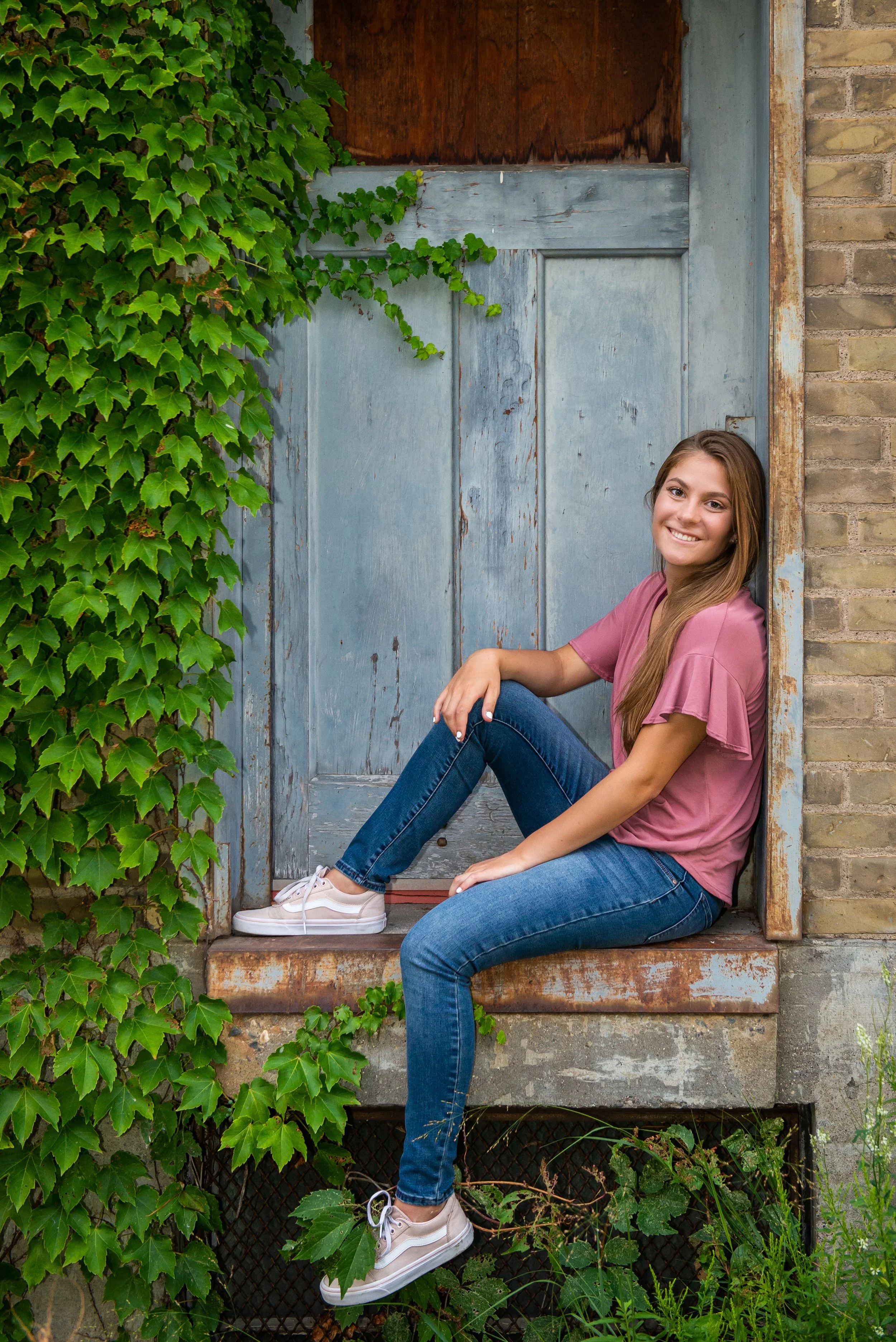 A young woman with long brown hair, wearing a pink top, blue jeans, and white sneakers, sitting on the doorstep of a rustic building with a weathered blue door and ivy growing on the side, smiling at the camera.