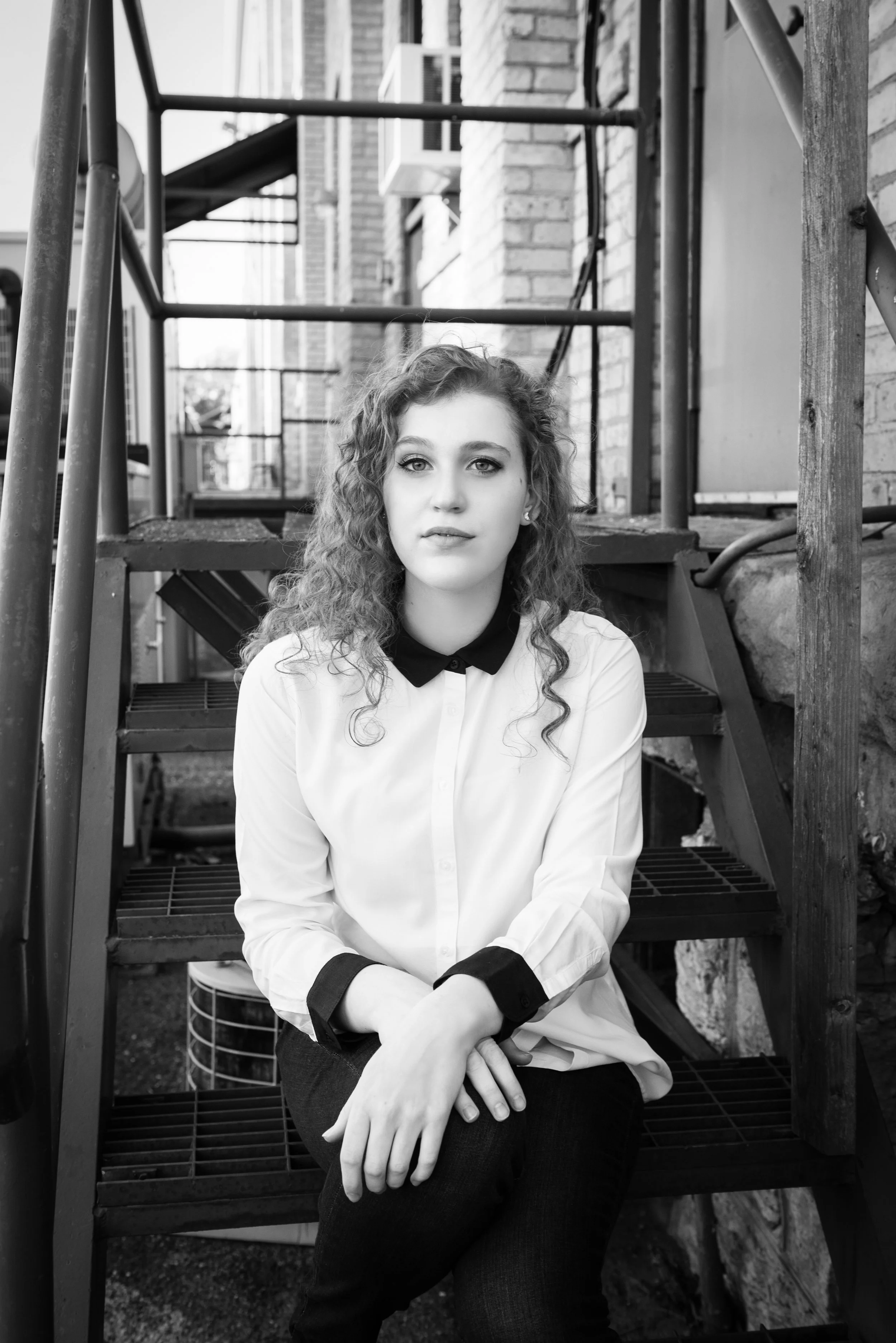 A young woman with curly hair and fair skin sitting on outdoor stairs, wearing a white shirt with black collar and cuffs, looking into the camera with a neutral expression.