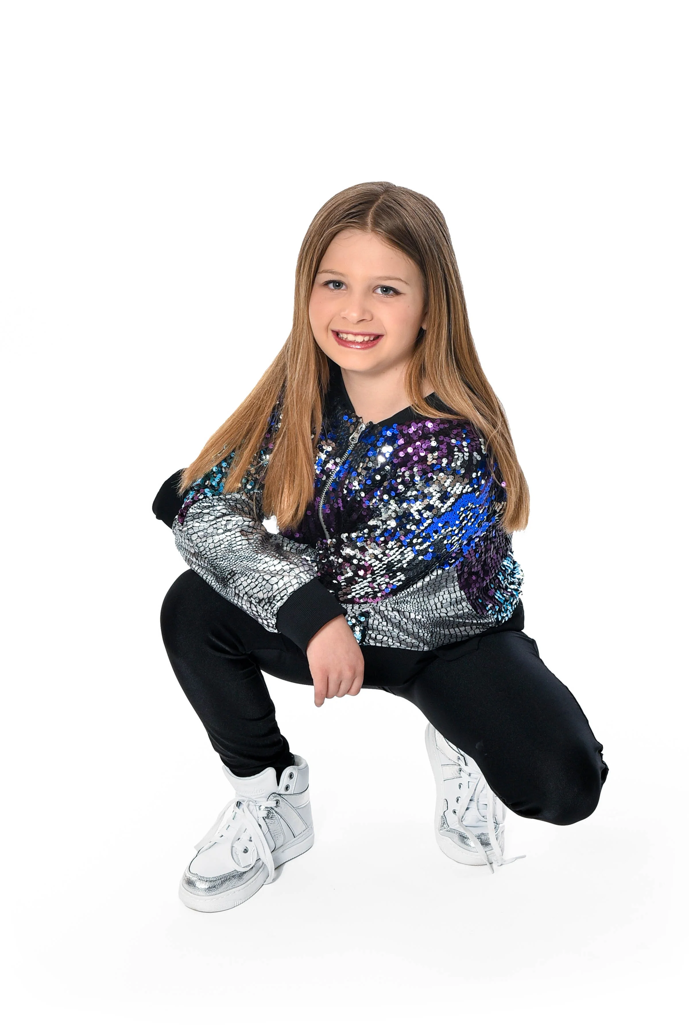 A young girl with long, light brown hair squatting and smiling, wearing a black, silver, purple, and blue sequined jacket, black pants, and white high-top sneakers.