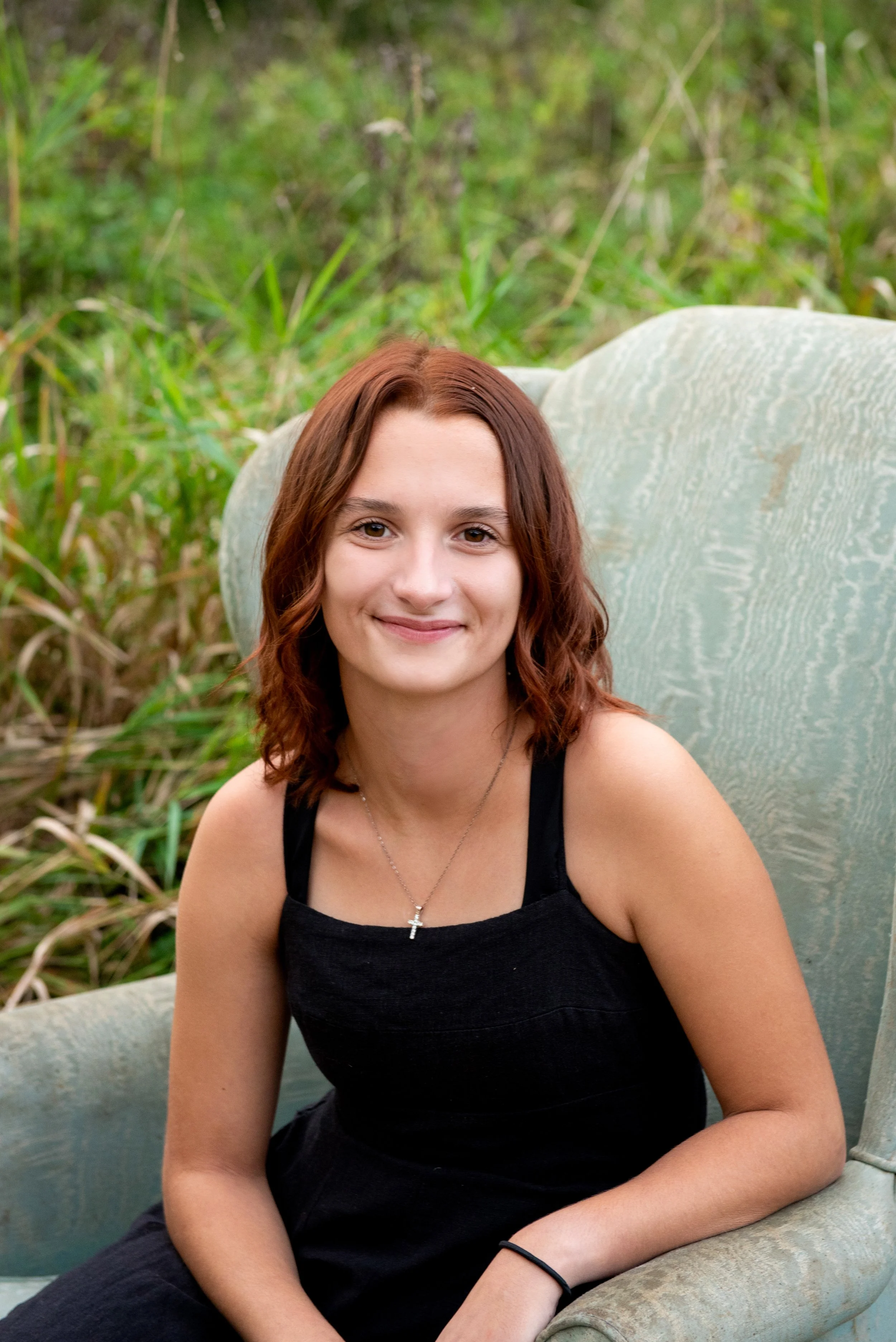 A young woman with shoulder-length auburn hair, wearing a black tank top and a cross necklace, sitting outdoors on a light green vintage armchair, with green foliage in the background.