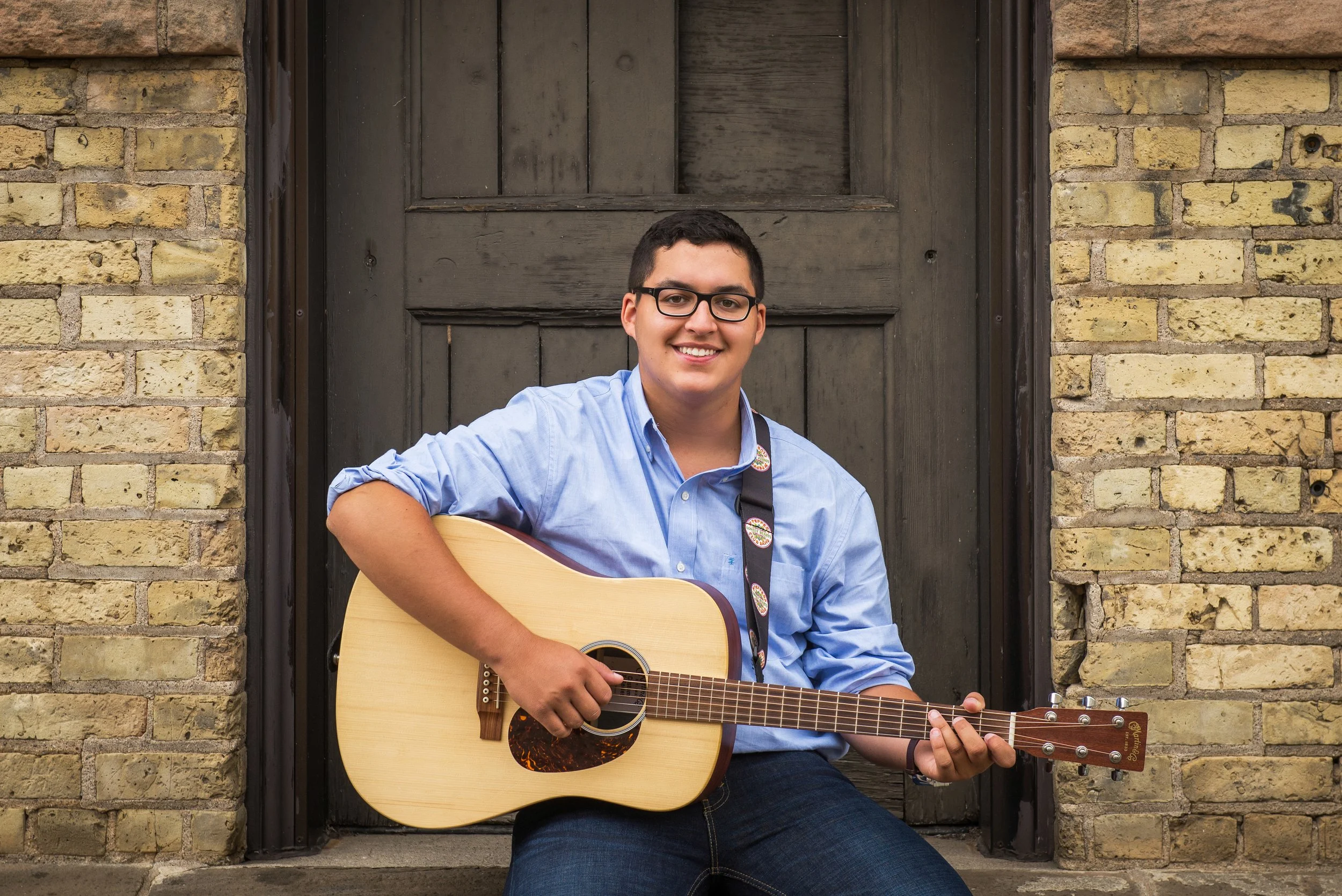 Young man with glasses sitting on a doorstep, playing an acoustic guitar, against a brick and wooden door background.