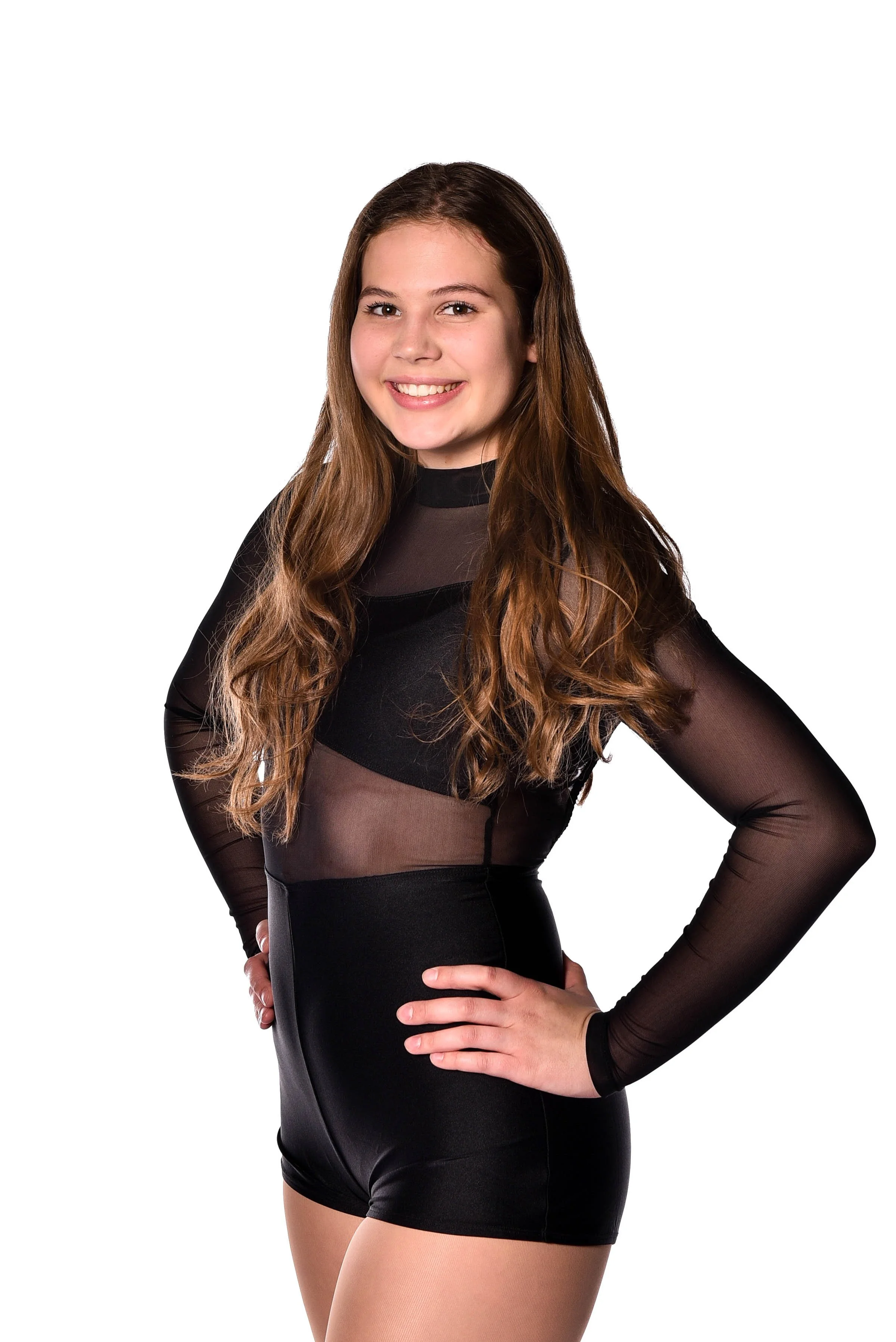A young woman with long brown hair smiling, wearing a black sheer long-sleeve top and black shorts, standing with one hand on her hip against a white background.