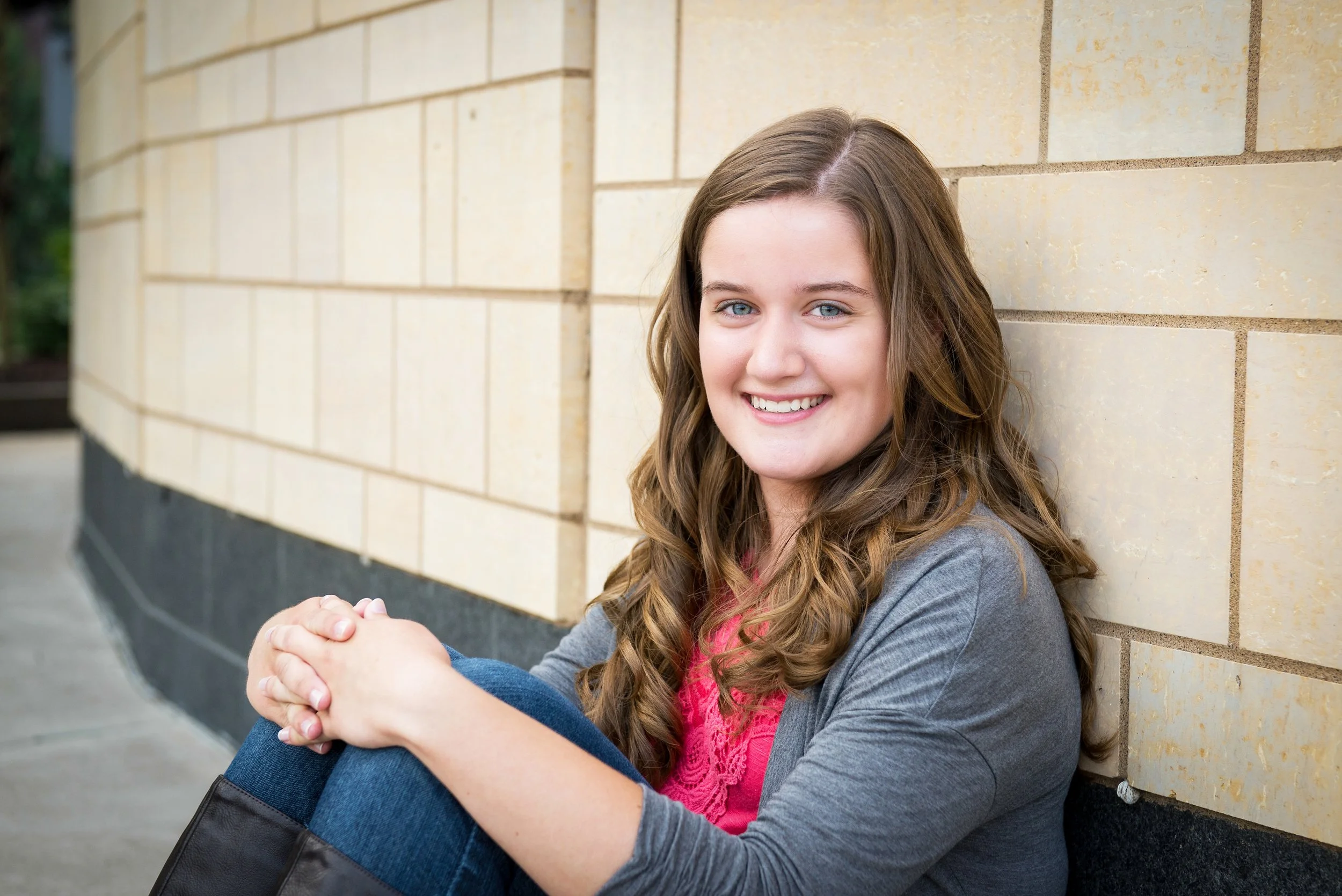 A young woman with long brown hair and blue eyes smiling while sitting against a beige brick wall outdoors.