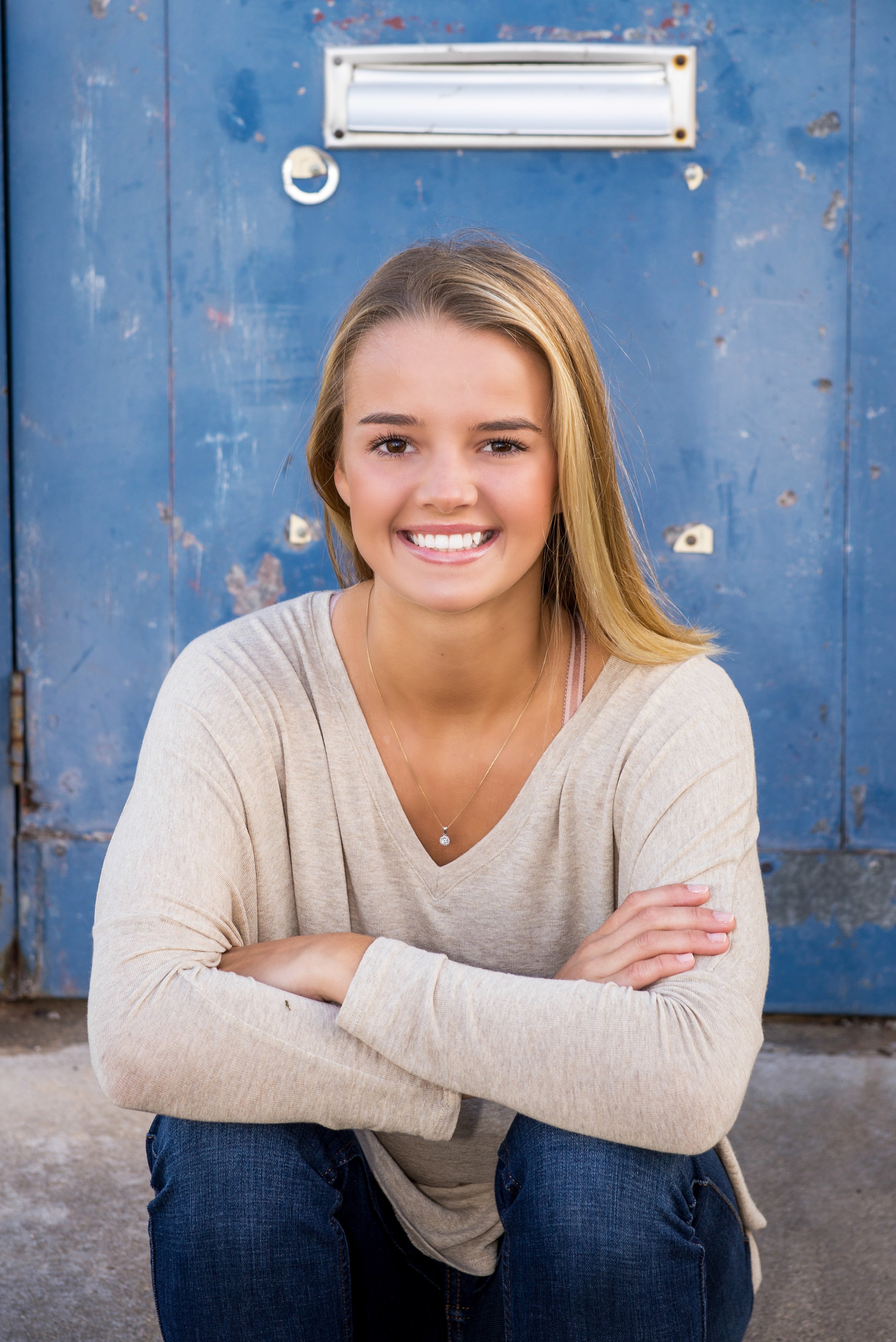 A young woman with long blonde hair, smiling, squatting in front of a blue metal locker. She is wearing a beige long sleeve shirt and blue jeans.