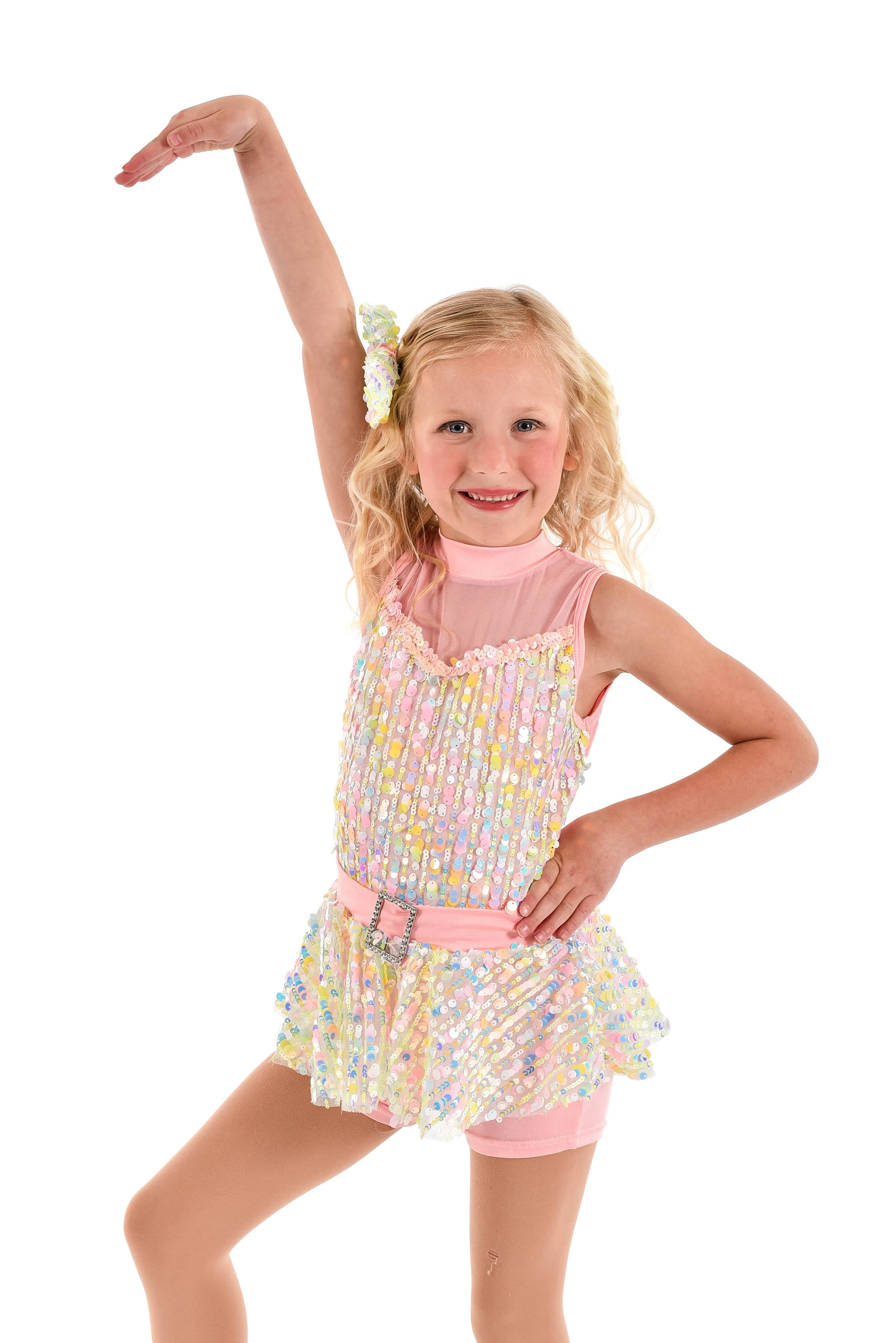 A young girl with curly blonde hair, smiling, wearing a pink sequin dress with a sheer pink neckline, a pink belt with a buckle, and pink shorts, posing with one arm raised and the other on her hip.