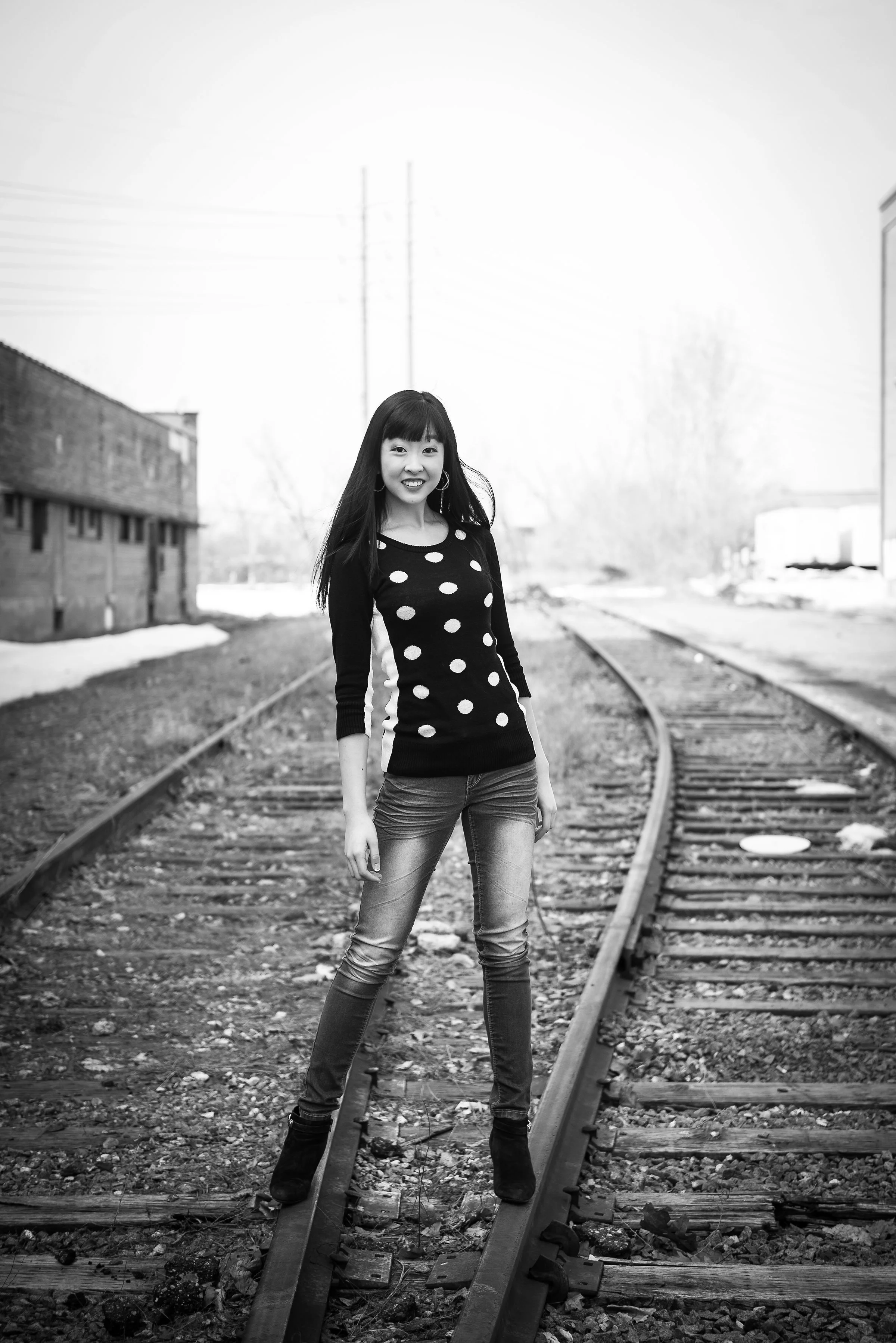 A young woman standing on a railway track outdoors, smiling at the camera, dressed in a polka dot sweater, jeans, and boots, in black and white.