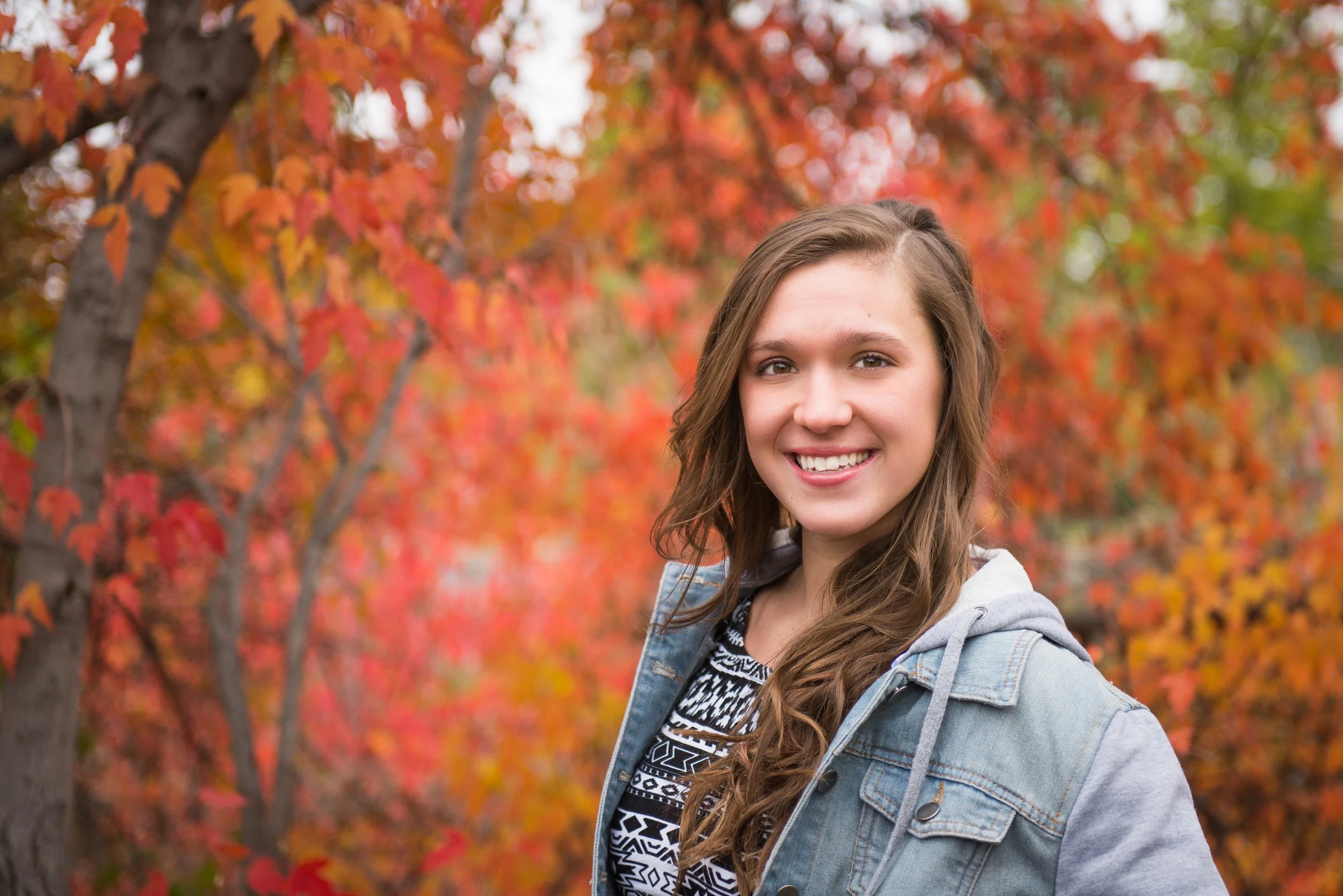 A smiling young woman with long wavy hair, wearing a patterned shirt and denim jacket, standing outdoors in front of vibrant red, orange, and yellow fall foliage.
