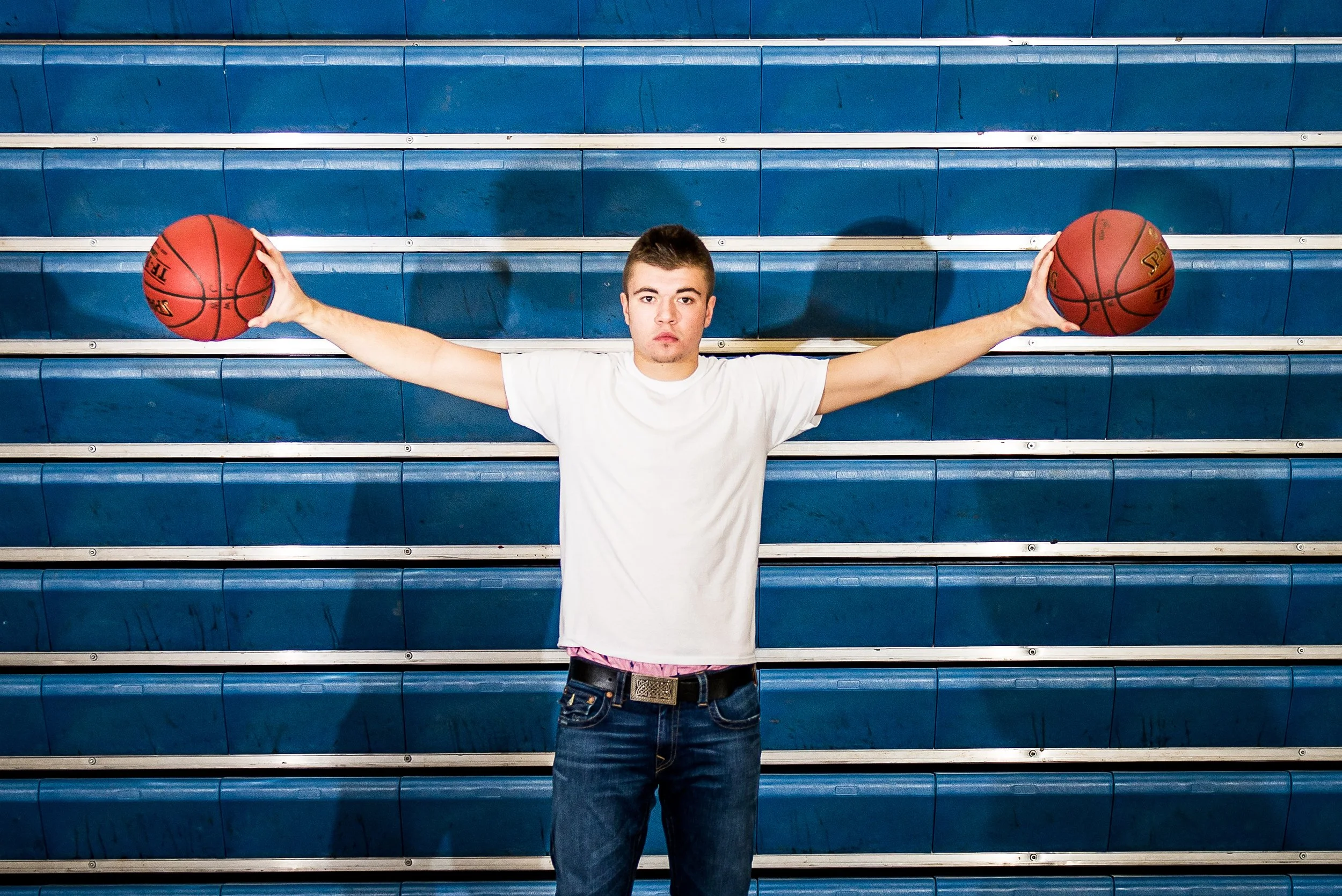 Young man standing in front of blue bleachers, holding a basketball in each hand with arms extended outward.
