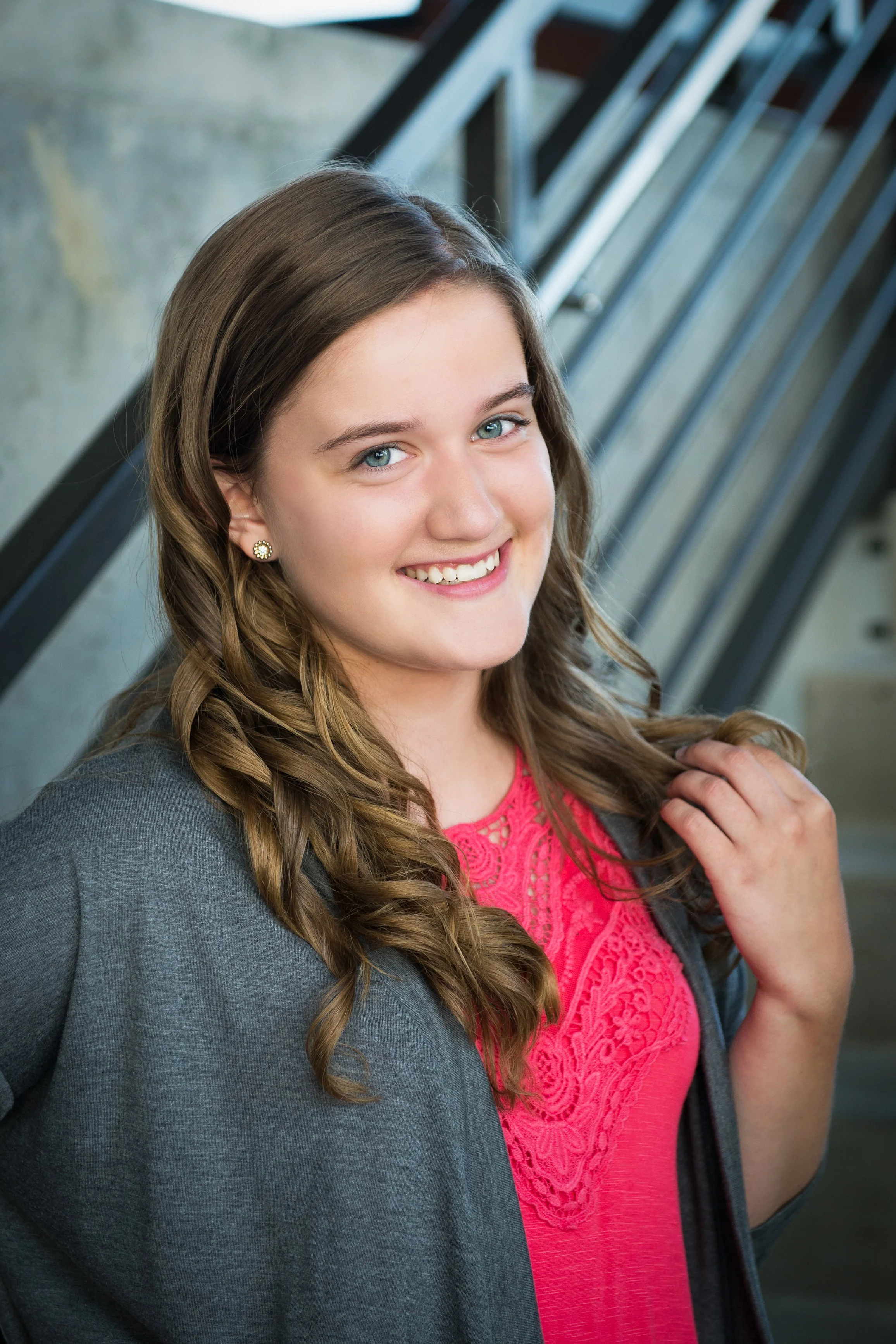 Young woman with long wavy brown hair, wearing a gray blazer and a pink lace top, smiling and standing near a staircase with metal railing.