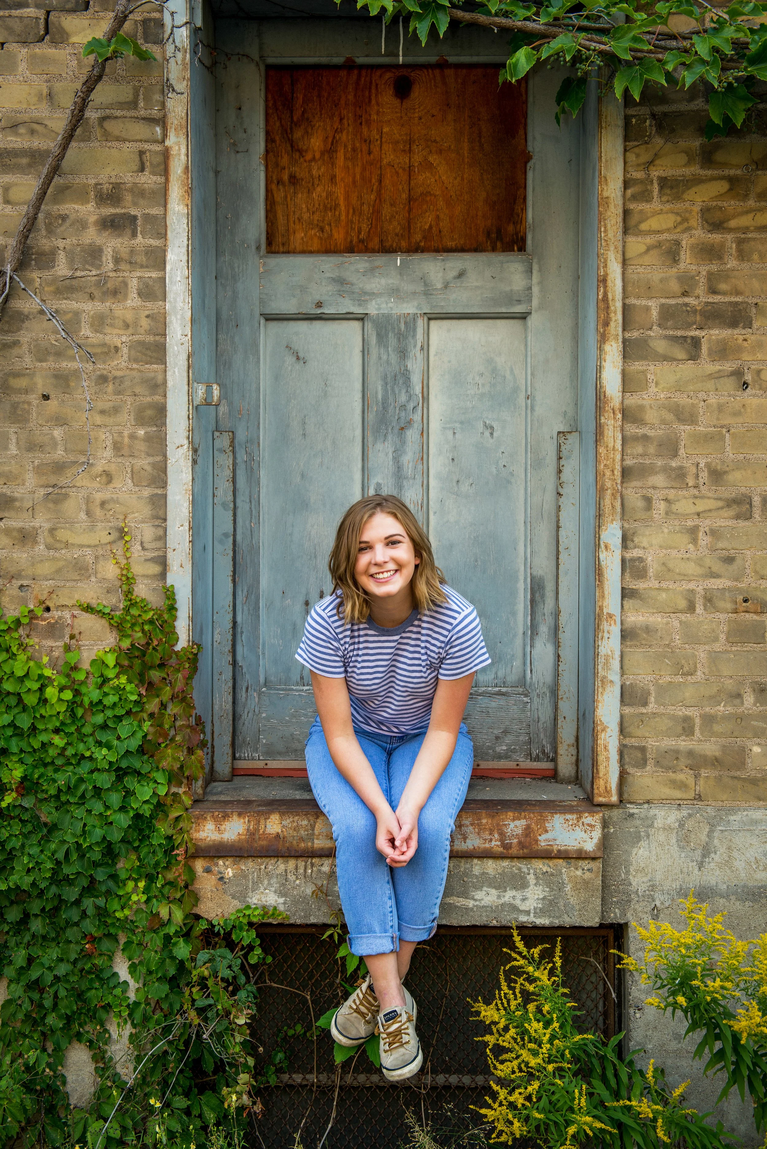 A young woman with shoulder-length brown hair, smiling, sitting on a step in front of a weathered, blue wooden door set into a brick wall, surrounded by greenery.