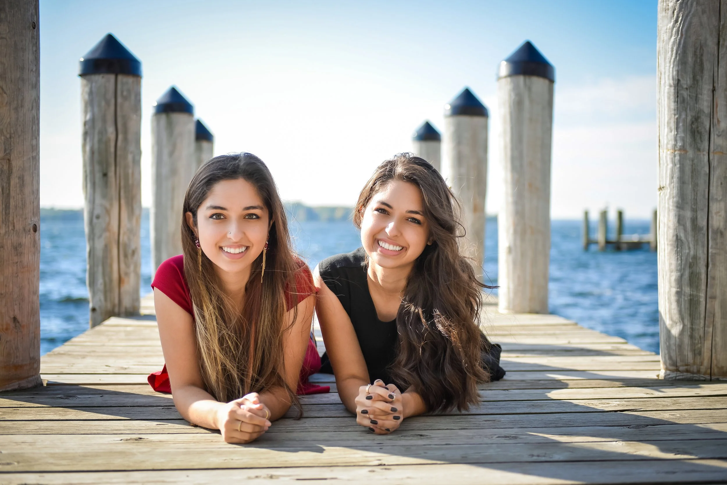 Two young women with long hair lying on a wooden dock by the water, smiling at the camera, with a blue sky and water in the background.