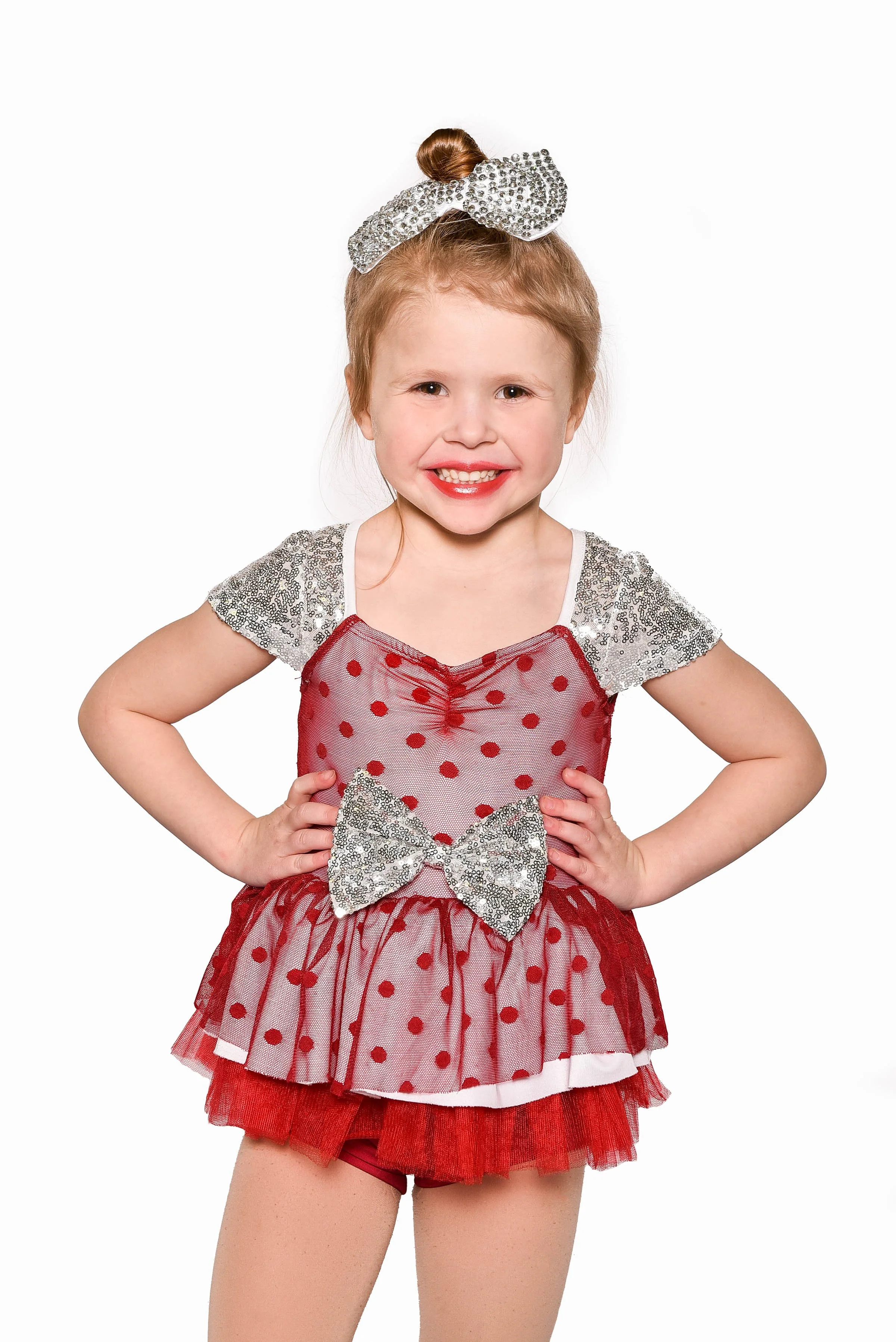 Young girl with red hair in a bun, wearing a red and silver polka dot dress with a bow and tutu, smiling with hands on hips, against a white background.