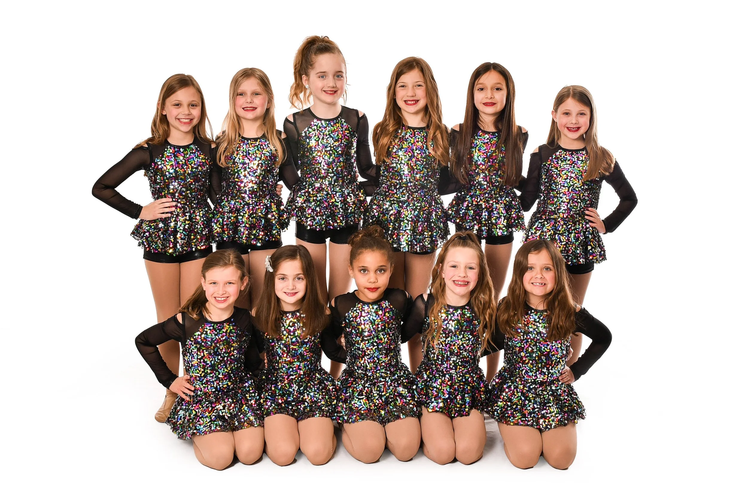 A group of twelve young girls dressed in matching black and multicolored sequin costumes, posing together for a photo against a white background.