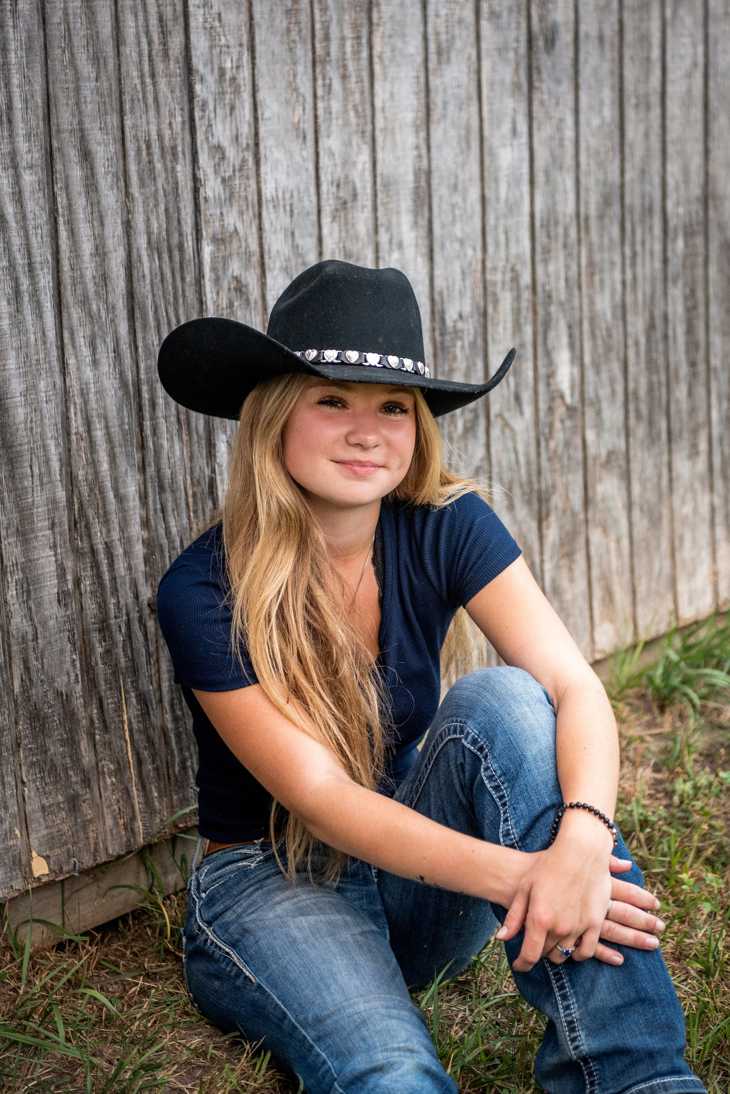 A young woman with long blonde hair sitting on the ground against a wooden fence, wearing a cowboy hat, a navy blue T-shirt, and jeans.