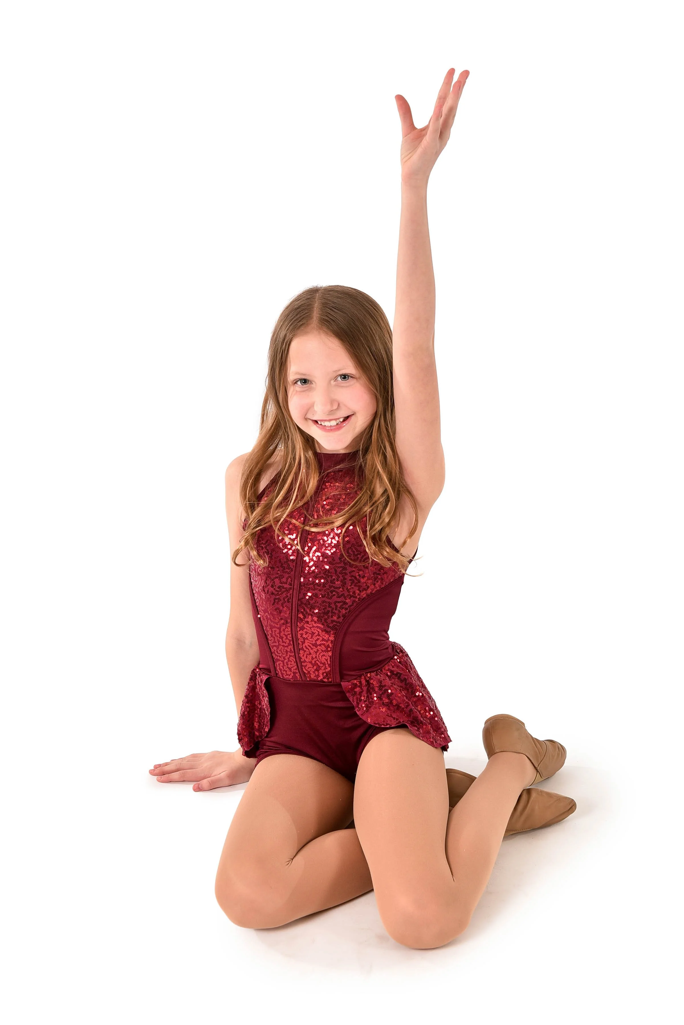 Young girl in a red, sequined dance costume, kneeling on the floor with one arm raised, smiling, against a plain white background.