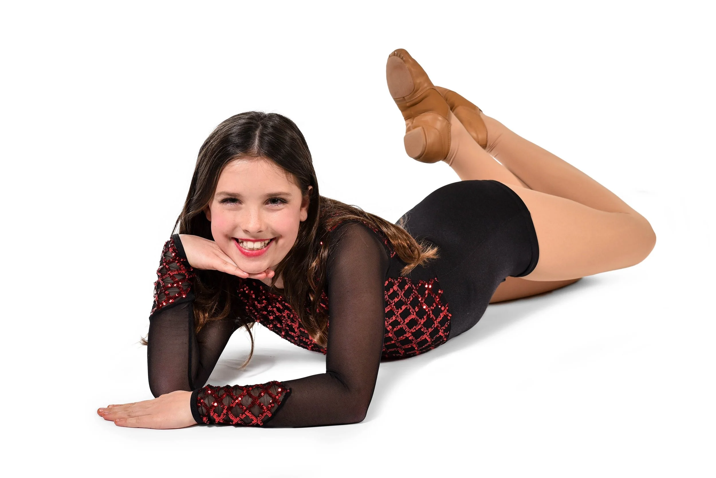 Young woman lying on her stomach with chin resting on hand, smiling, wearing a black and red dance costume and tan tights, against a white background.