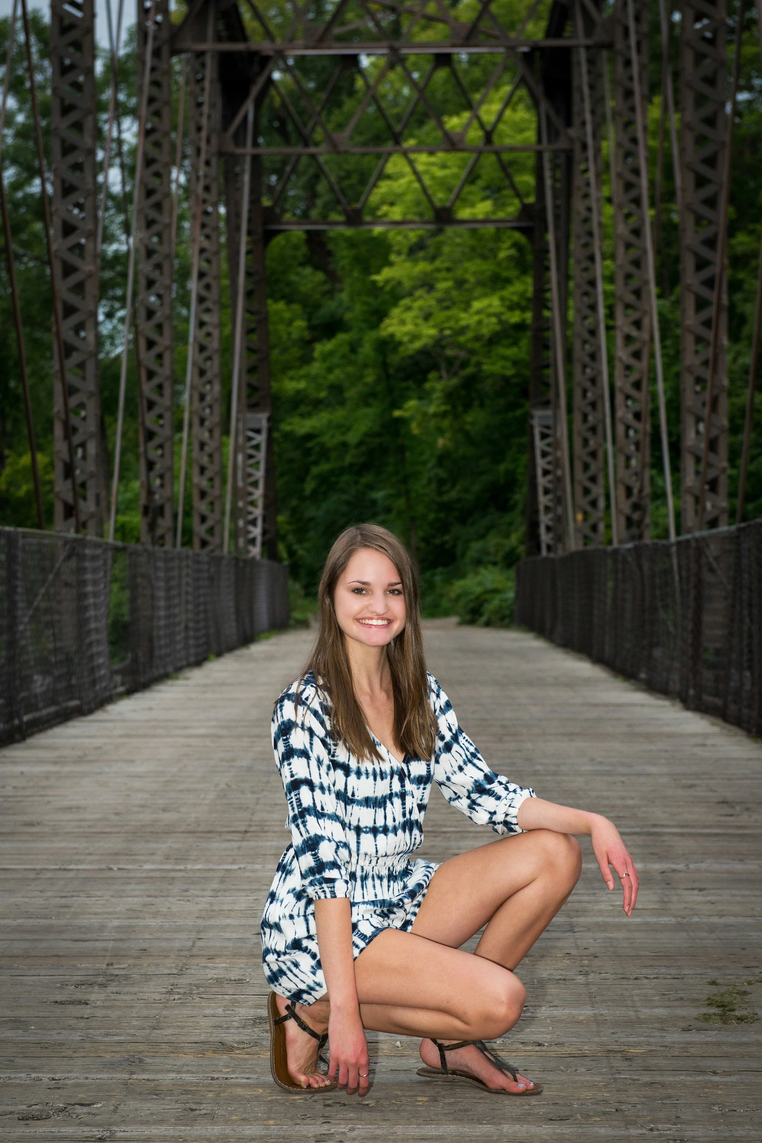 A young woman with long brown hair, smiling, is crouching on a wooden bridge during daytime. She is wearing a black and white tie-dye dress and sandals. The bridge has black railings and a metal truss structure above, with green trees in the backgrou