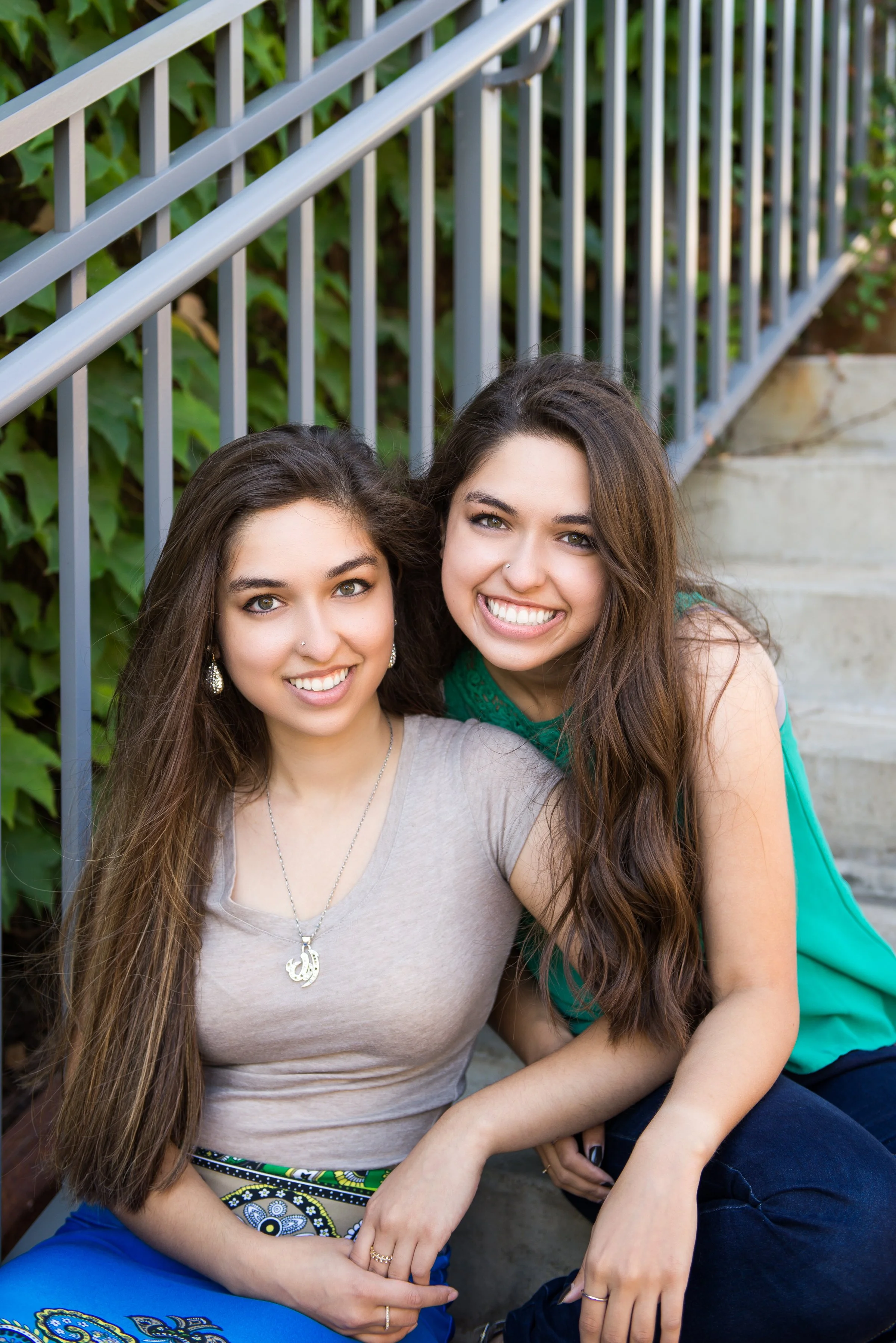 Two young women sitting on outdoor stairs near a metal railing, smiling at the camera, with greenery in the background.