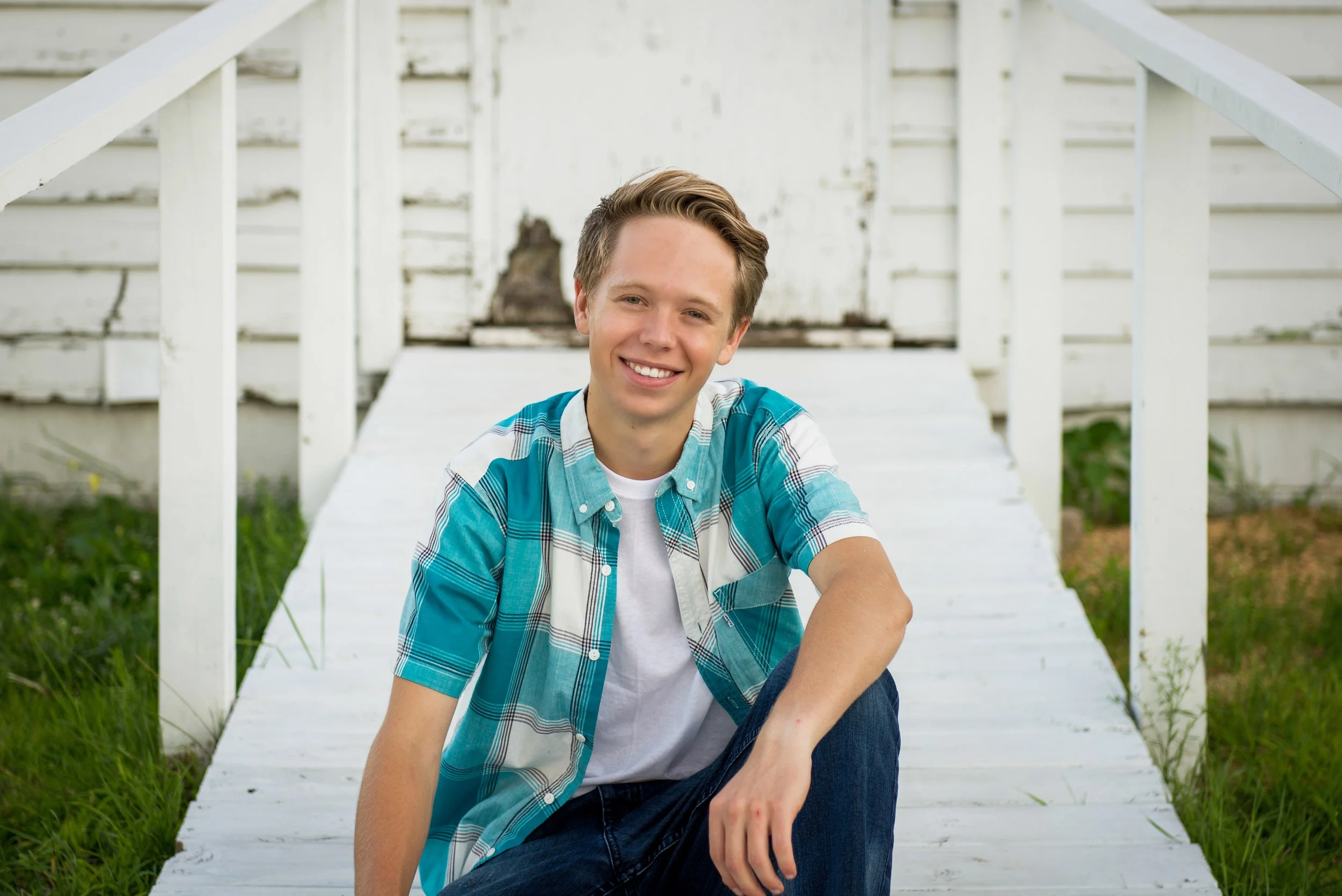 A young man with short brown hair smiling, sitting on a white wooden walkway leading to an old white house with a staircase, wearing a blue plaid shirt over a white t-shirt.
