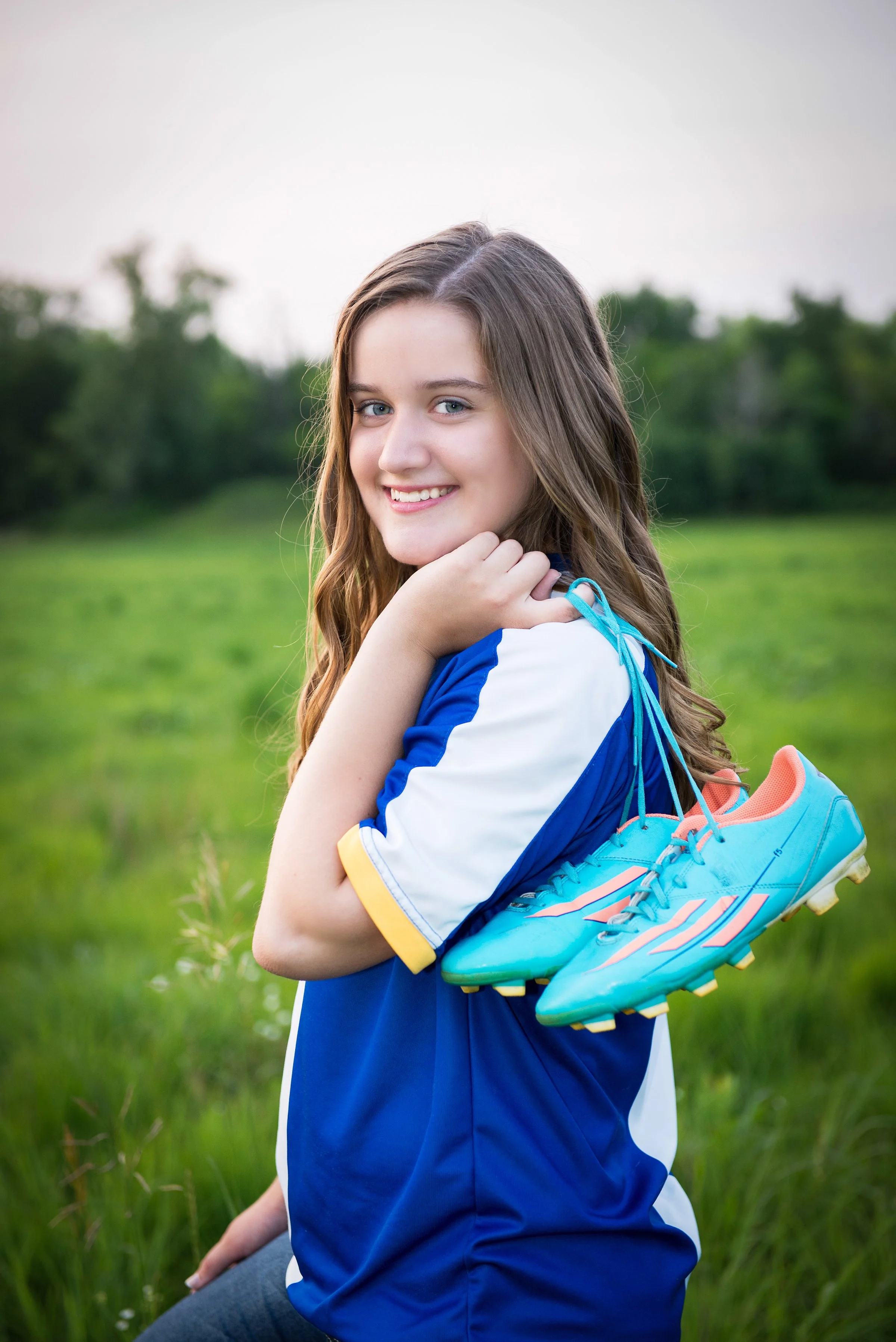 A young woman with long, wavy brown hair and blue eyes smiling while holding a pair of blue and pink soccer cleats over her shoulder. She is standing outdoors in a grassy field with trees in the background.