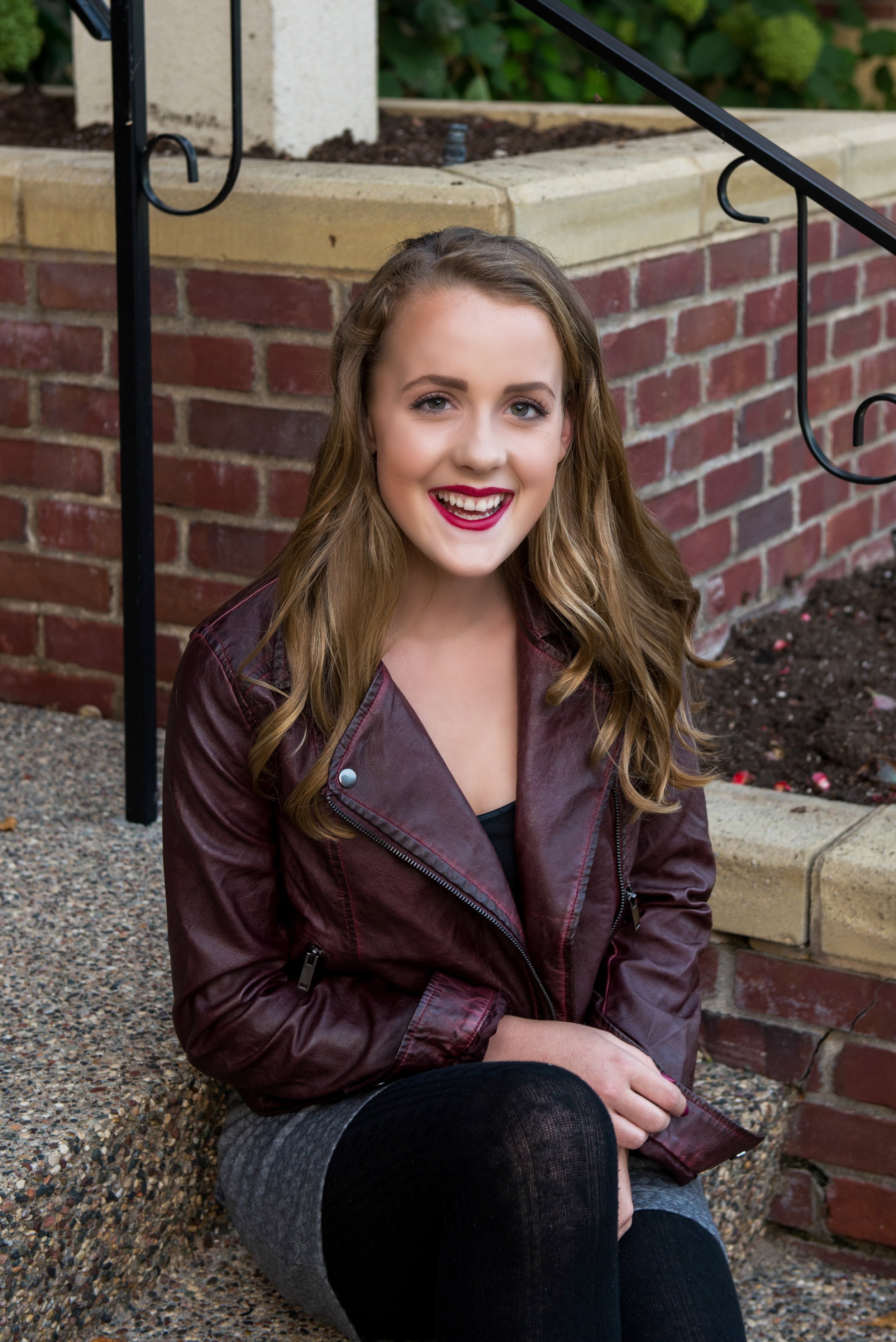 Young woman with long wavy blonde hair sitting outside on a concrete step, smiling at the camera, wearing a maroon leather jacket, black top, gray skirt, and black tights, with a brick building and plant bed in the background.