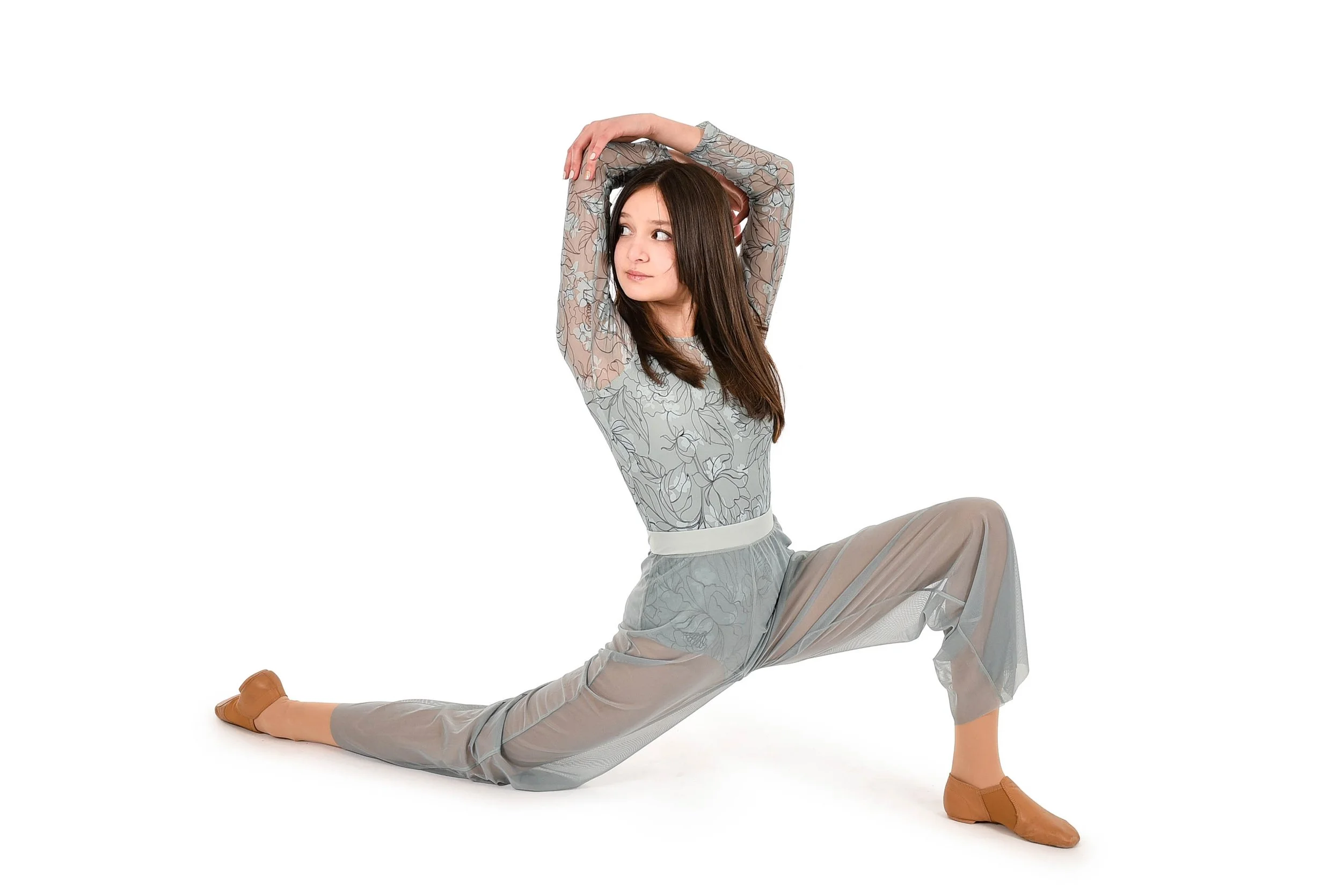 A young woman in gray floral-patterned clothing and tan dance shoes performing a split stretch with arms raised above her head against a white background.