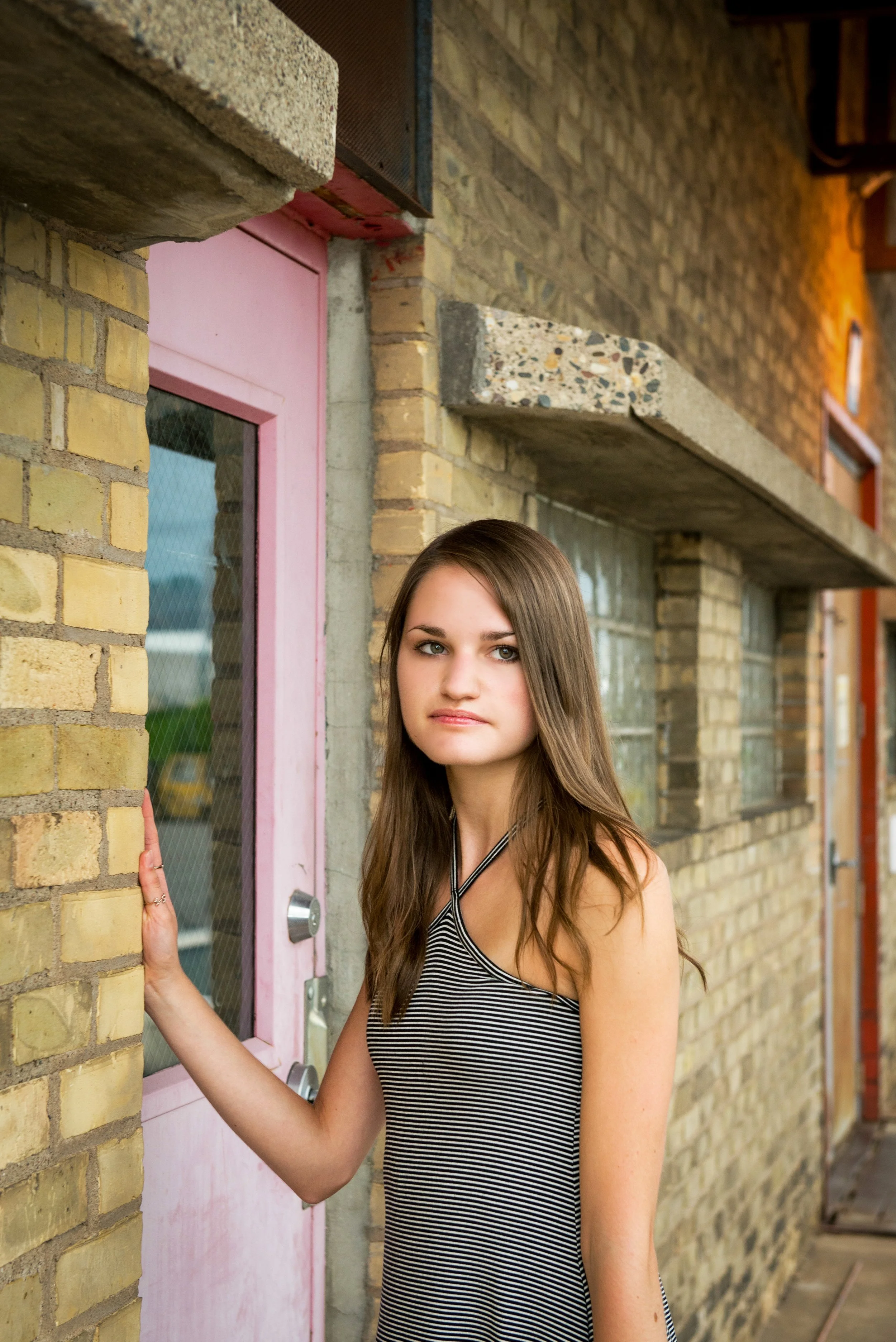 A young woman with long brown hair wearing a black and white striped sleeveless top stands near a pink door with a glass window, touching the brick wall with her left hand, in an outdoor urban setting.