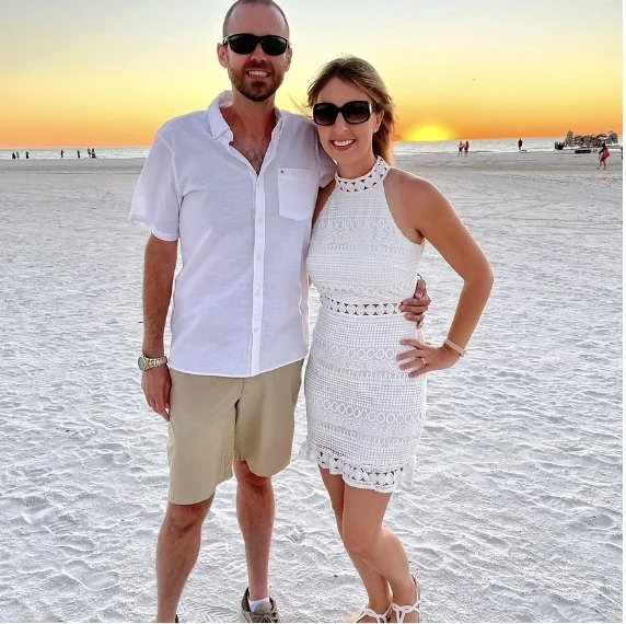 A man and woman standing on a beach at sunset, smiling and posing for a photo.