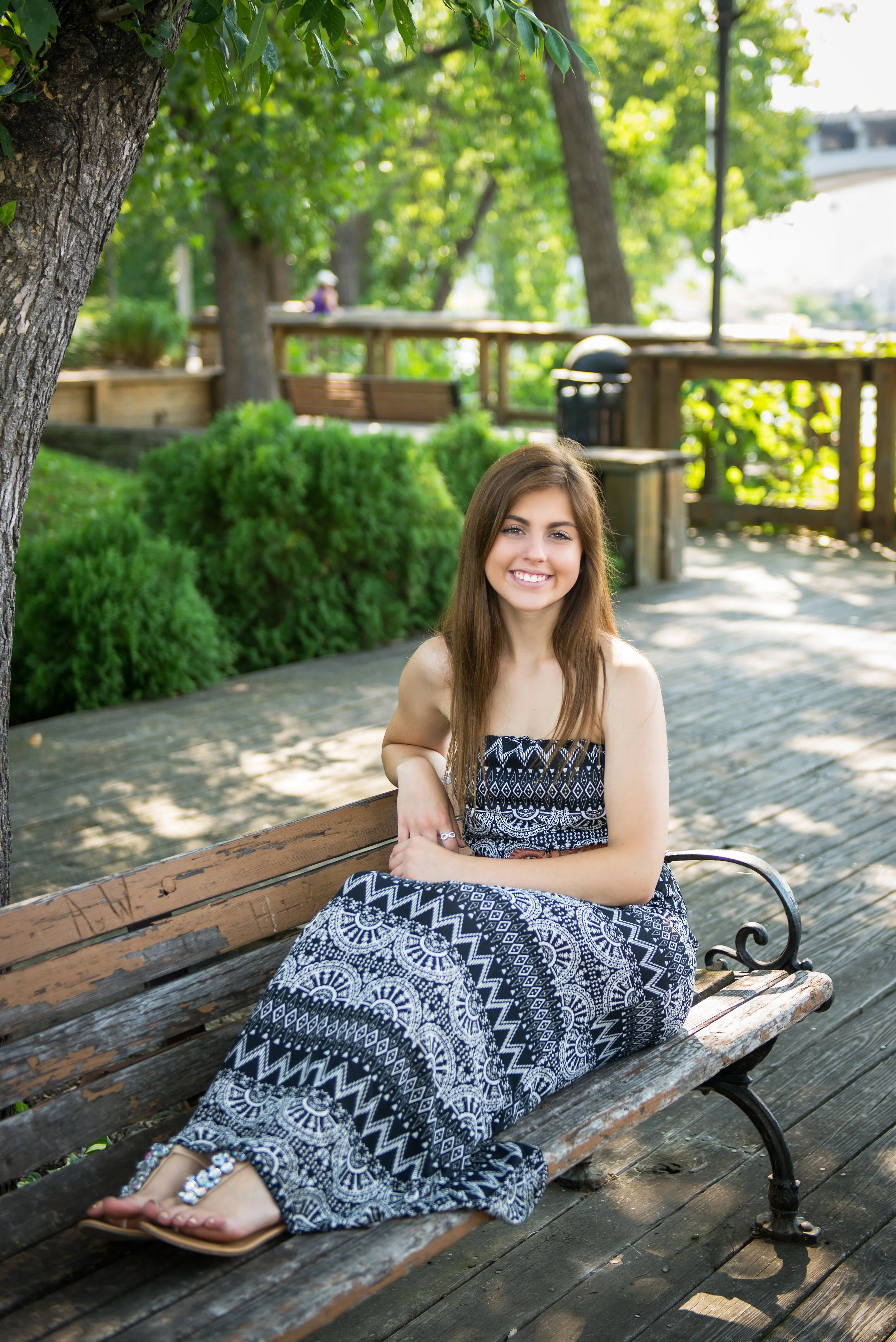 A young woman in a patterned strapless maxi dress with long brown hair, sitting on a wooden park bench under a tree, smiling at the camera with greenery and a walking path in the background.