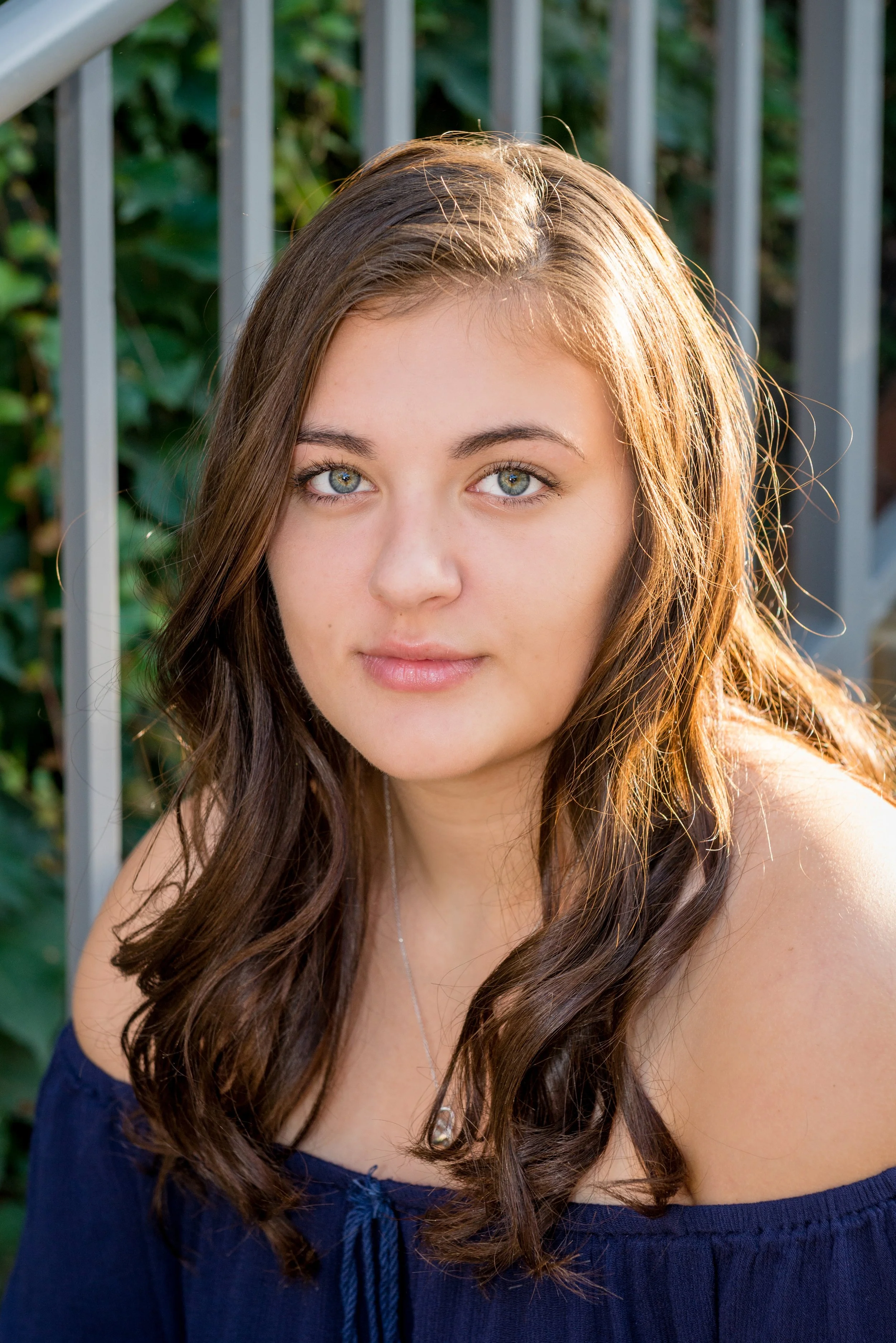 A young woman with long wavy brown hair and blue eyes, wearing an off-the-shoulder navy top, sitting outdoors near a metal fence with greenery in the background.