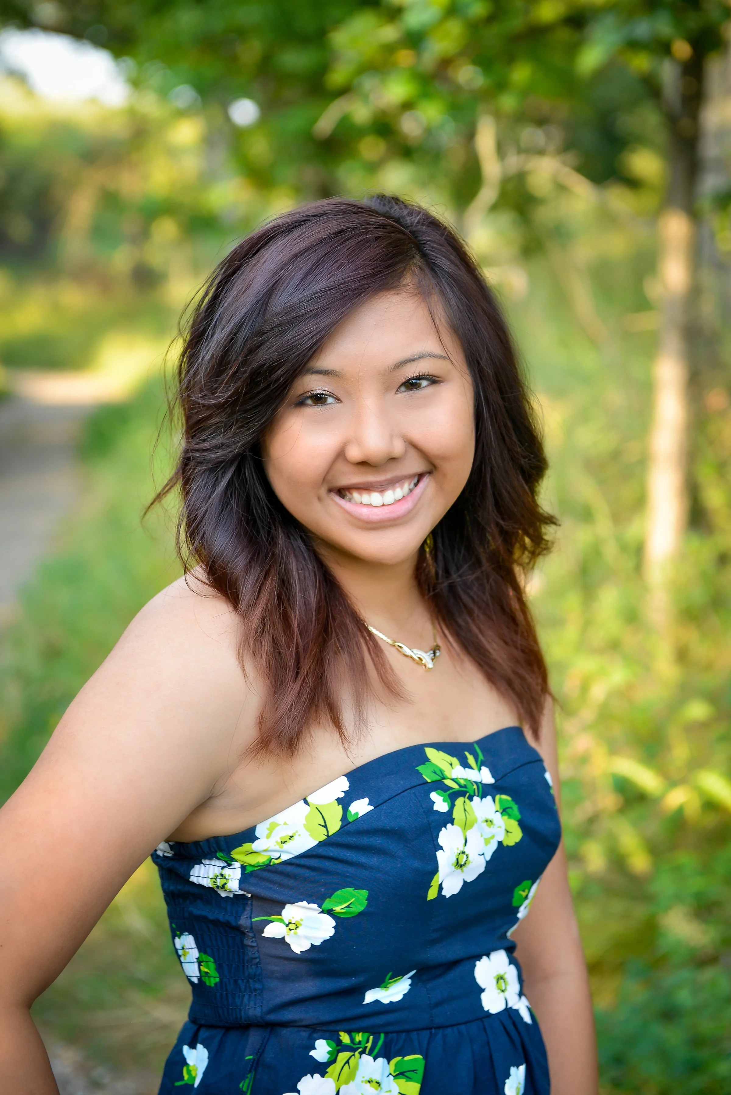 A young woman with shoulder-length dark hair and a bright smile, wearing a strapless blue dress with white and green floral patterns, standing outdoors surrounded by green trees and foliage.