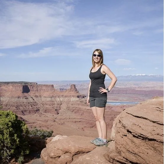 Woman standing on rock ledge at Grand Canyon with arms on hips, wearing sunglasses, black tank top, and shorts, smiling