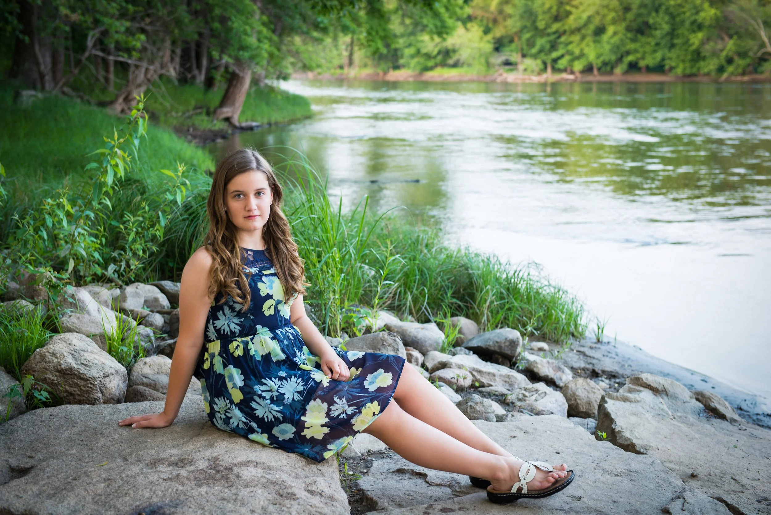 A young woman with long brown hair sitting on a large rock by a river, surrounded by green grass and trees.