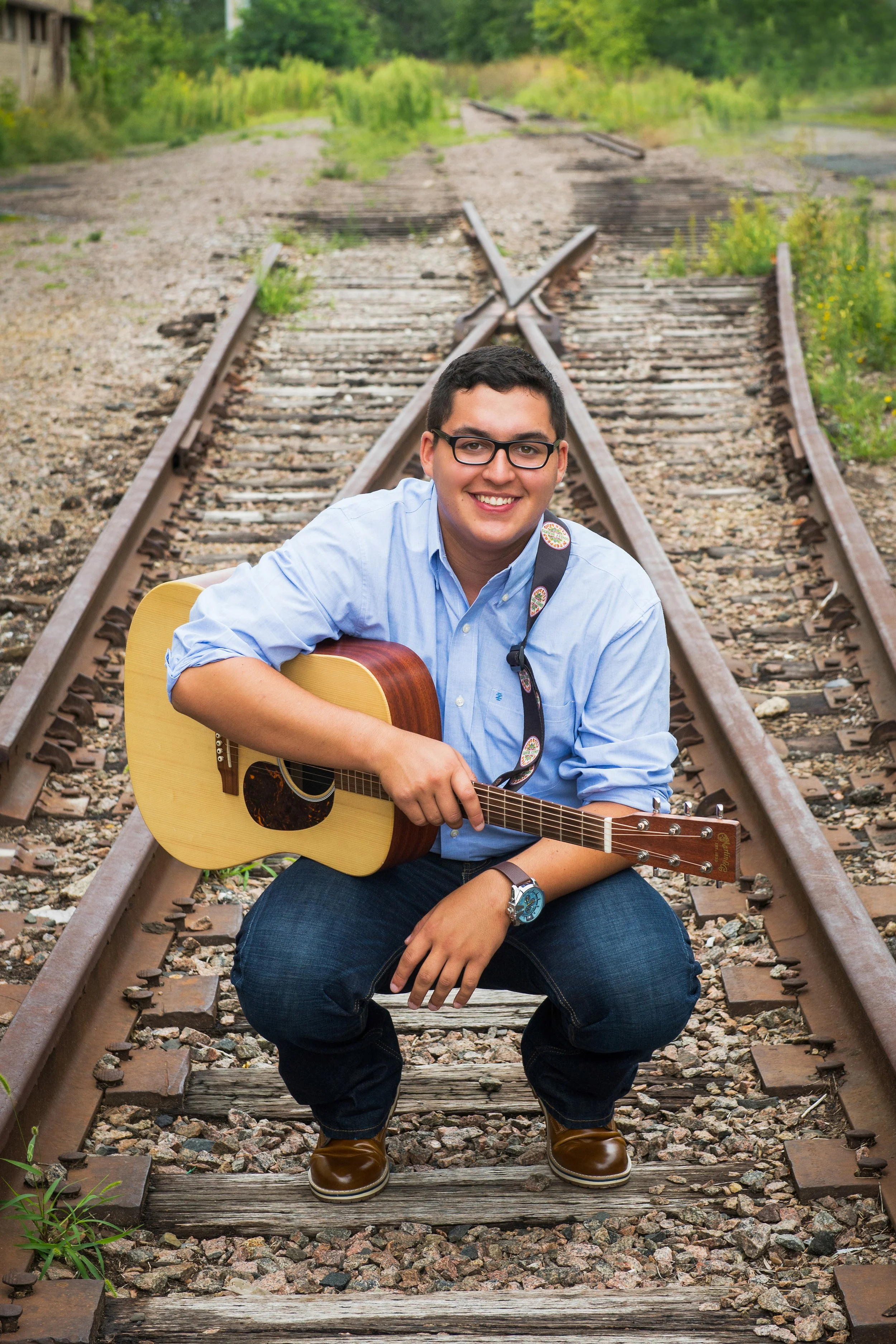 A young man with glasses smiling and sitting on old train tracks, holding an acoustic guitar, surrounded by greenery.