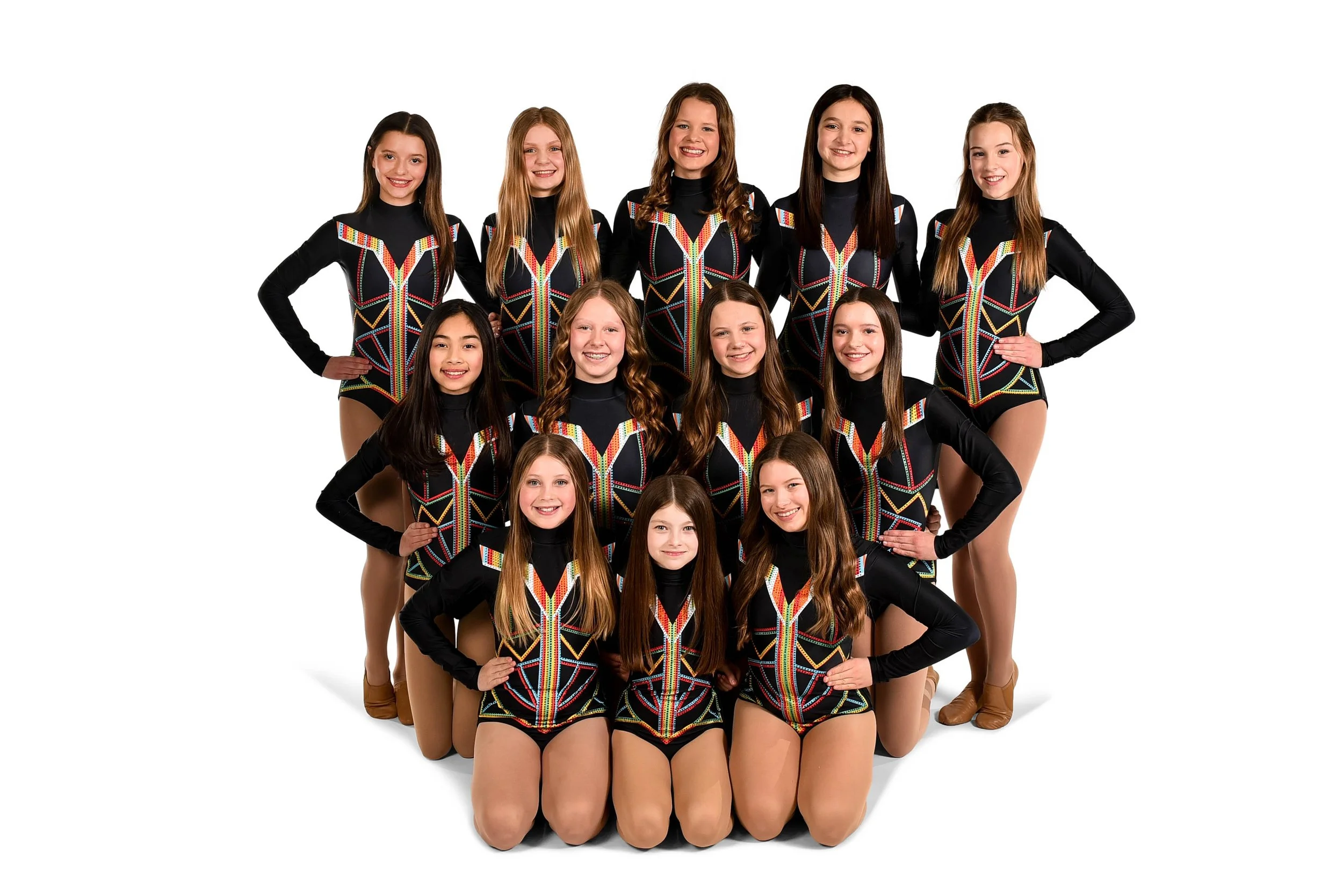 A group of fifteen young female figure skaters in matching black and colorful costumes, posed on the ice rink against a white background.