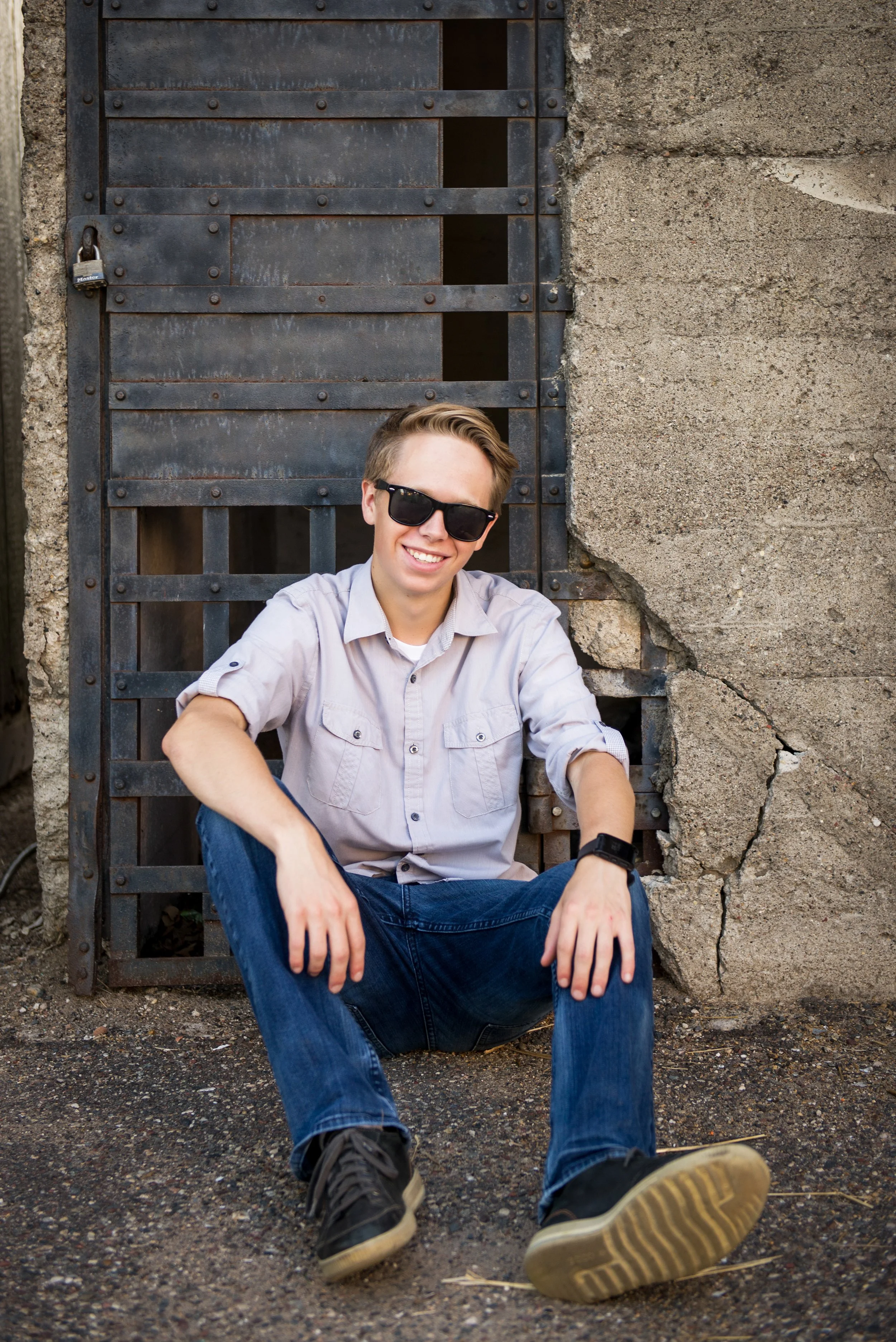 Young man in sunglasses, sitting on the ground, smiling, in front of a cracked, old concrete wall with a metal gate secured with a padlock.