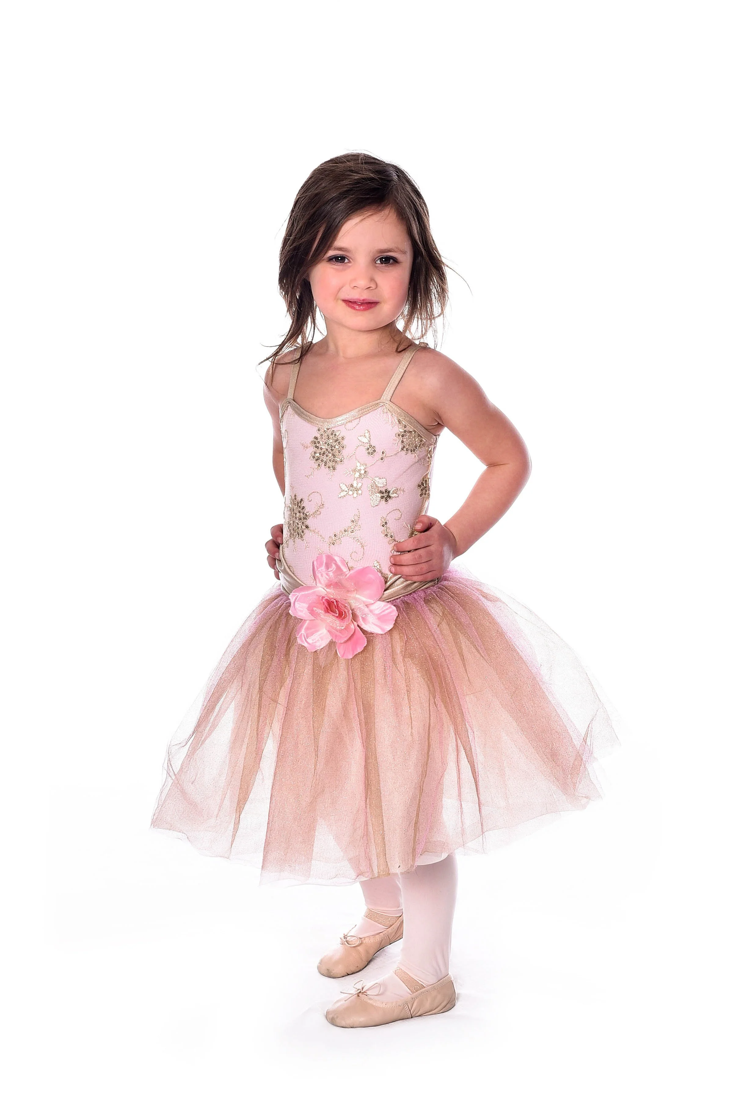Young girl in ballet costume with pink and gold tutu, floral embellishments, and ballet slippers, standing with hands on hips against a white background.