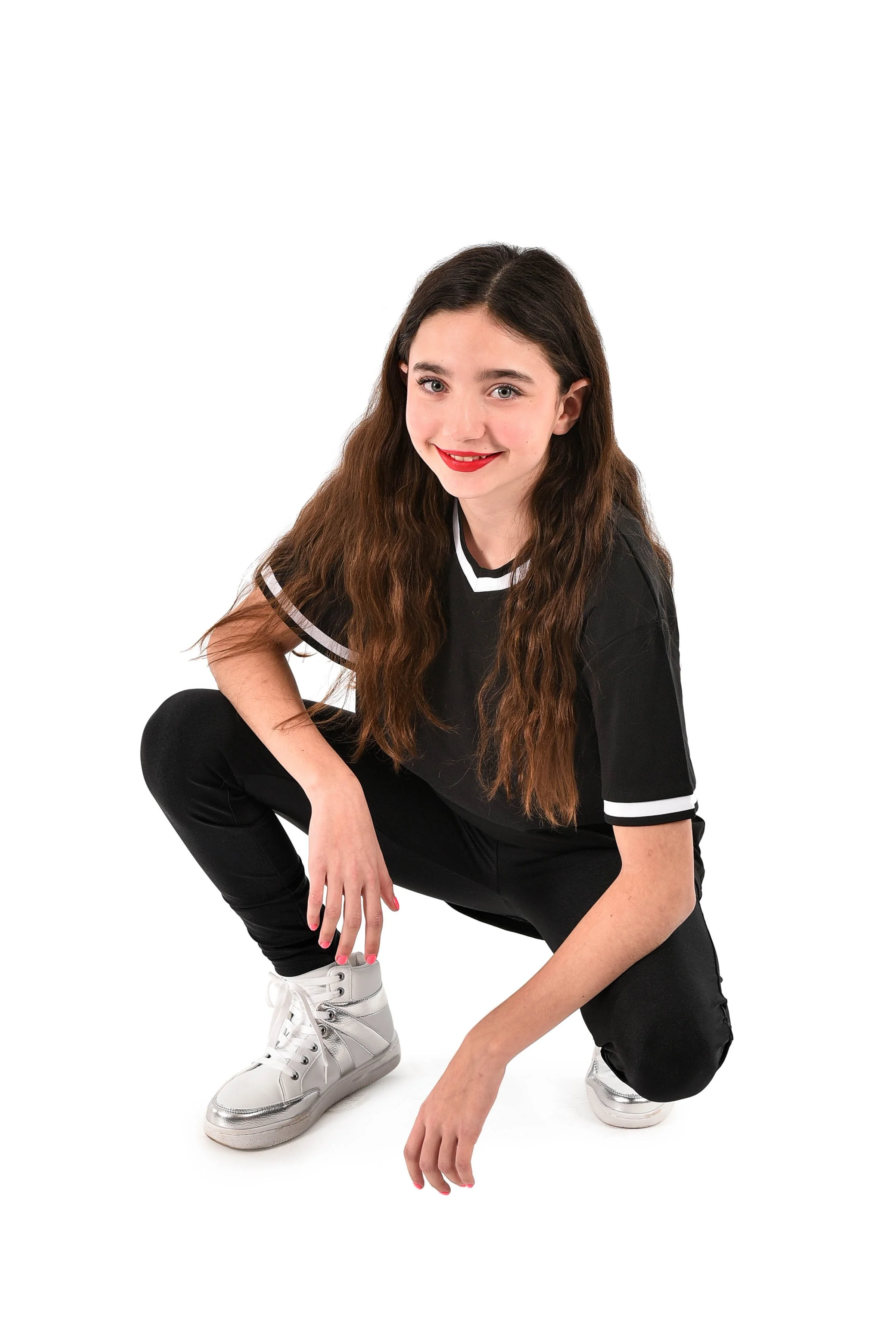 A young woman with long wavy brown hair crouching, wearing a black t-shirt with white trim, black pants, and white sneakers, smiling with red lipstick.