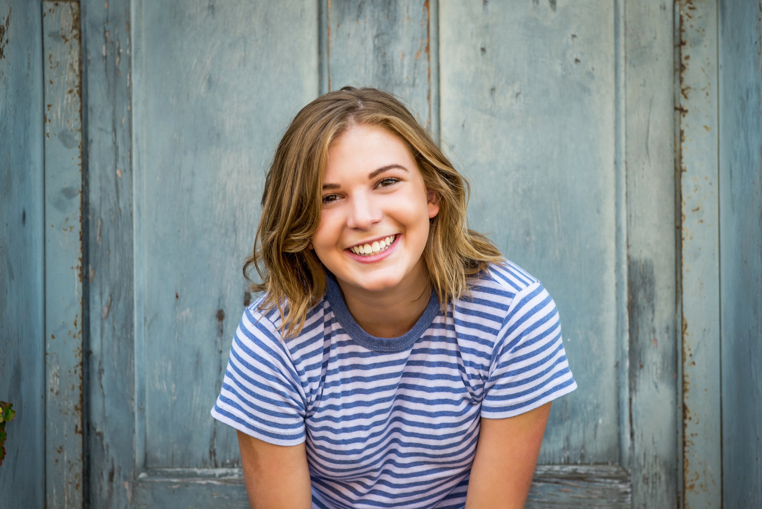 A young woman with shoulder-length light brown hair, wearing a blue and white striped t-shirt, smiling, against a weathered blue wooden background.